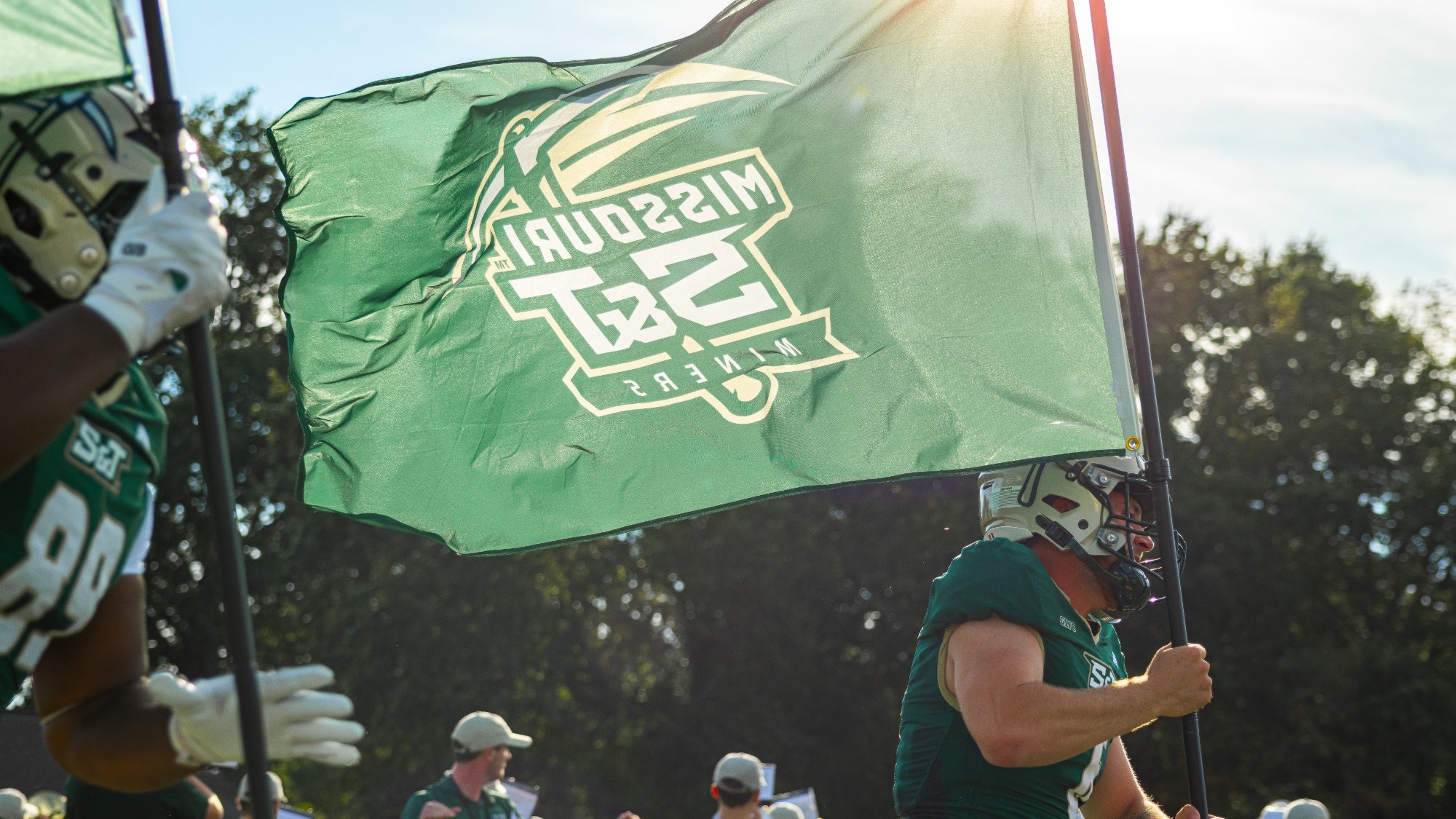 Eli Scott wearing a green uniform and gold helmet leading the Miners onto the field carrying a large green flag with the S&T logo on it