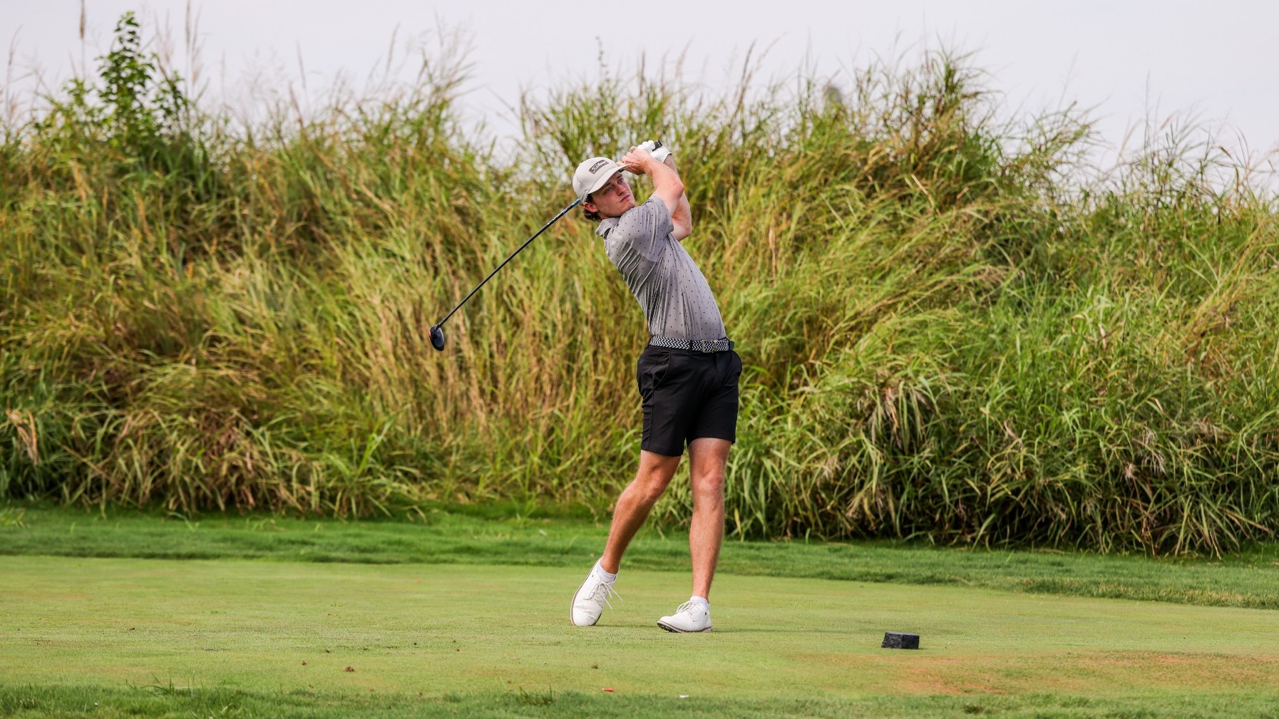 Thomas Heisner follows through on a golf swing on the tee box, wearing a gray polo, black shorts, white cap and white golf shoes, with tall grass lining the fairway behind him.