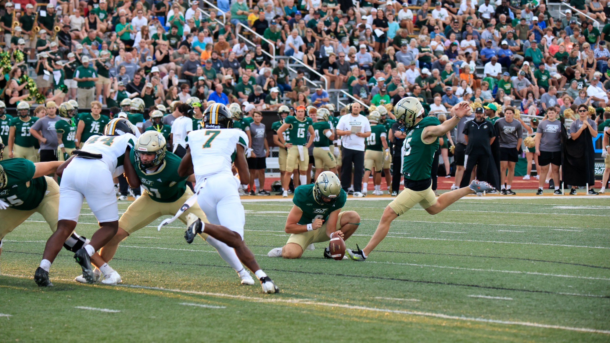 Layne Ryals photographed mid-kick during a football game at Allgood-Bailey Stadium while his teammate Logan Jacobson holds the ball for a field goal. They are both wearing green jerseys and gold helmets.