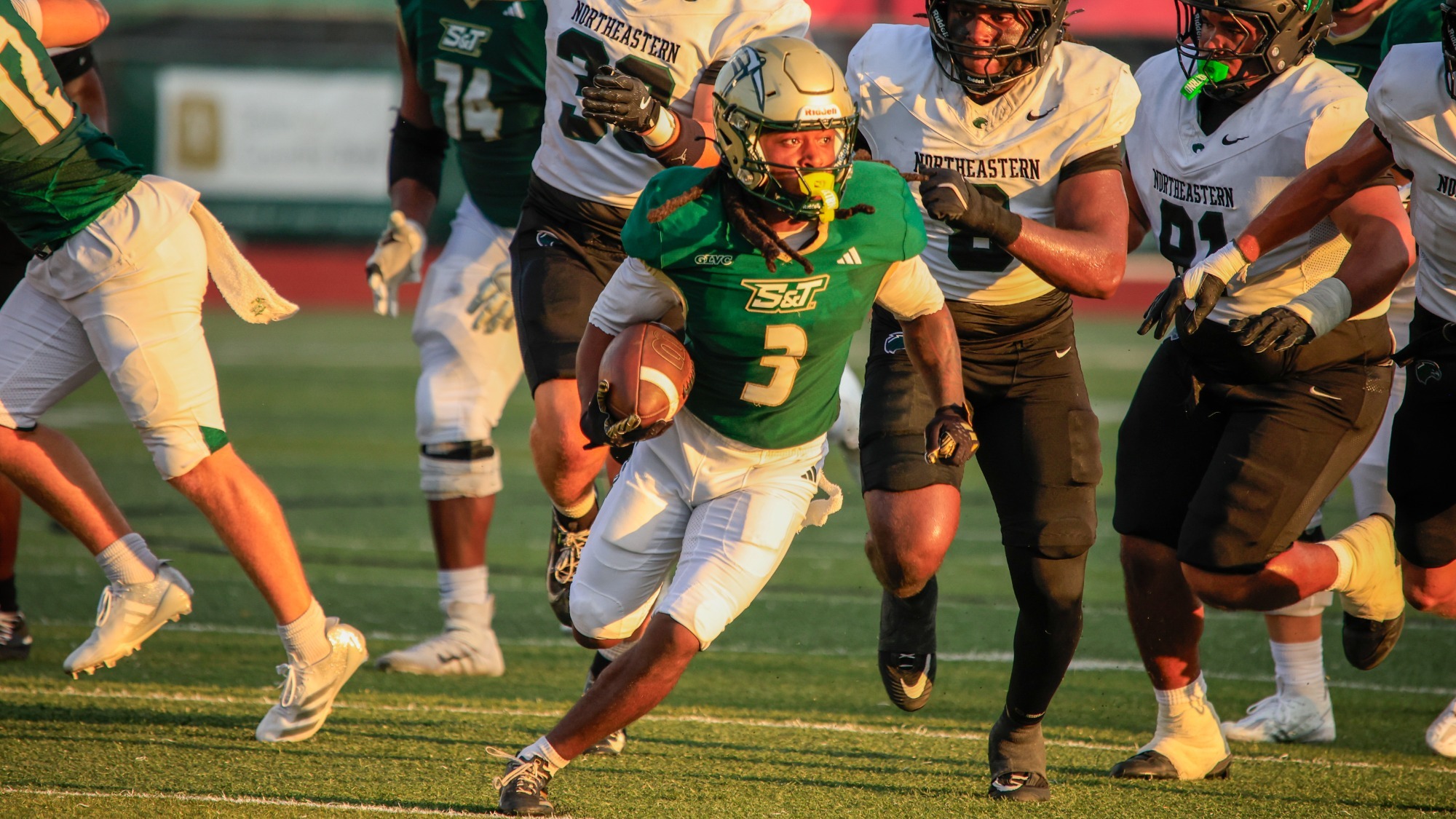 K'Vion Bills of Missouri S&T photographed running with the ball while wearing a green uniform and gold helmet, he is being chased by several NSU players wearing white and black