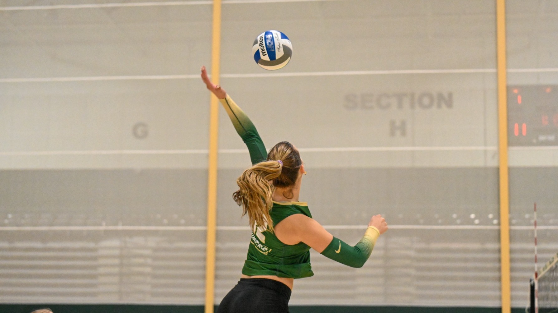 Dani Wilke of Missouri S&T women's volleyball photographed wearing a green uniform and jumping in the air and swinging her left arm at the volleyball as she attacks during a match at Missouri Southern