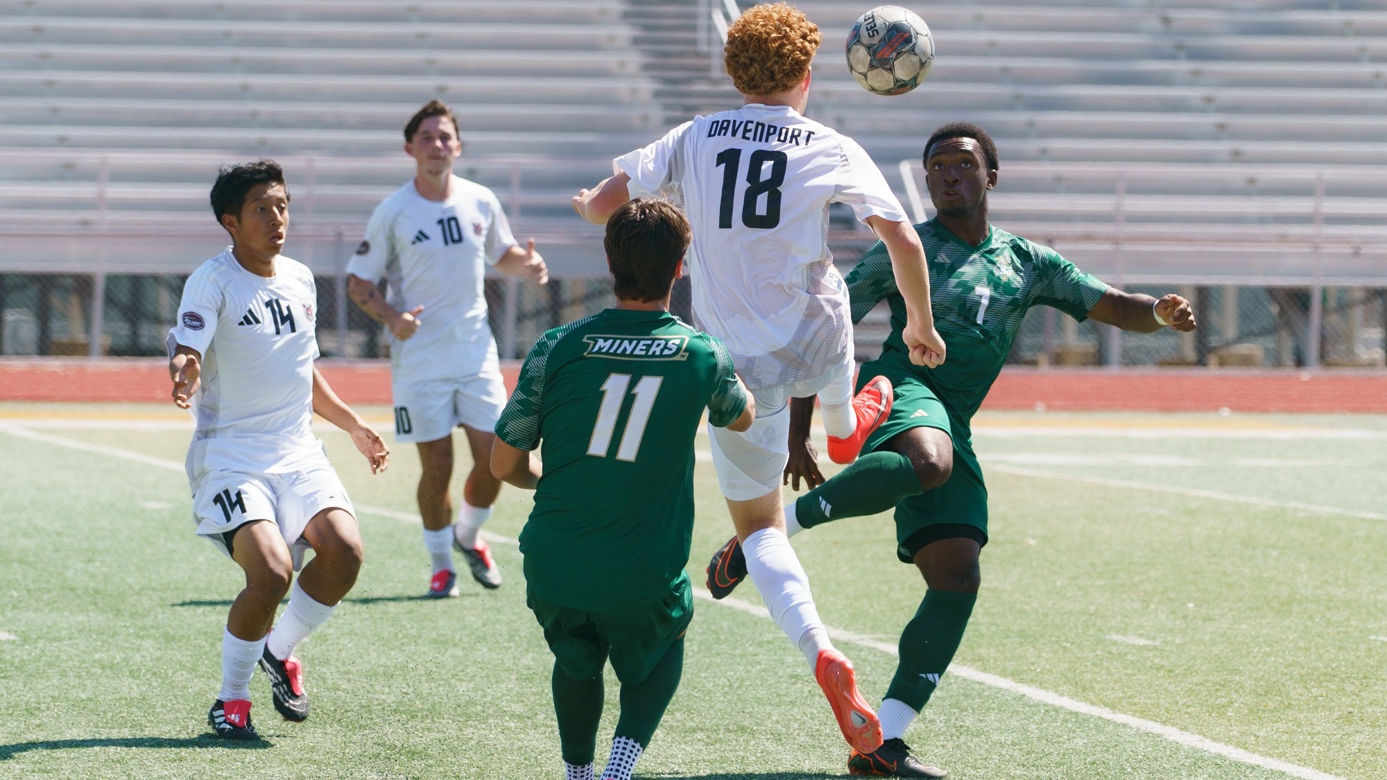 Khiba Romany of Missouri S&T Men's Soccer is wearing a green uniform and watching the ball as he goes to kick it out of the air with a Davenport defender in the foreground moving to play the ball too