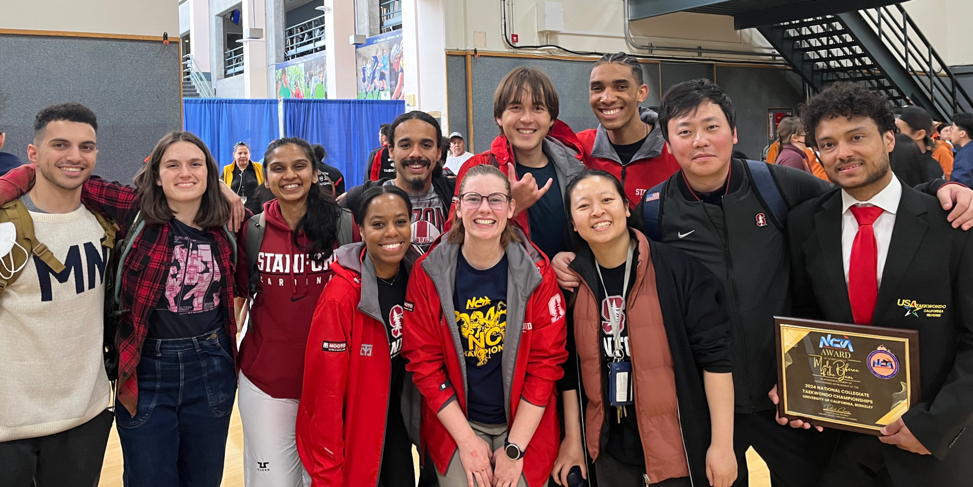 Photo shows several smiling members of taekwondo club wearing team apparel. Several members have medals. On the right, Coach Cristian is wearing a suit and holding an award. 