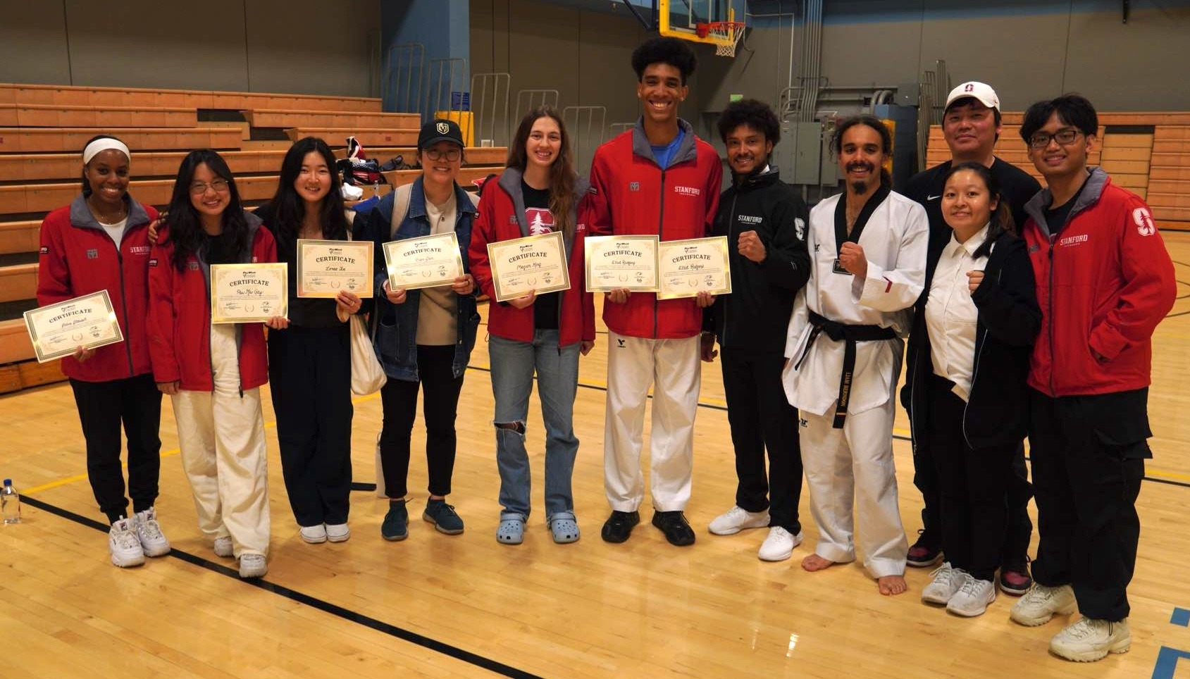 A group picture of 11 Taekwondo club members in a gymnasium. 6 of them are holding certificates indicating that they medaled at the event. 