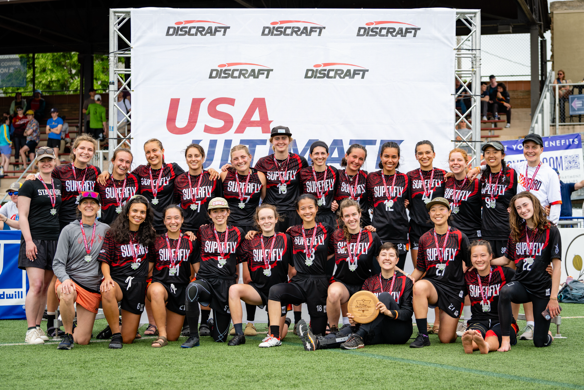 The Stanford women's ultimate team, Superfly, stands in front of a white USA Ultimate background in their dark jerseys after their championship game with silver medals around their necks, smiling.