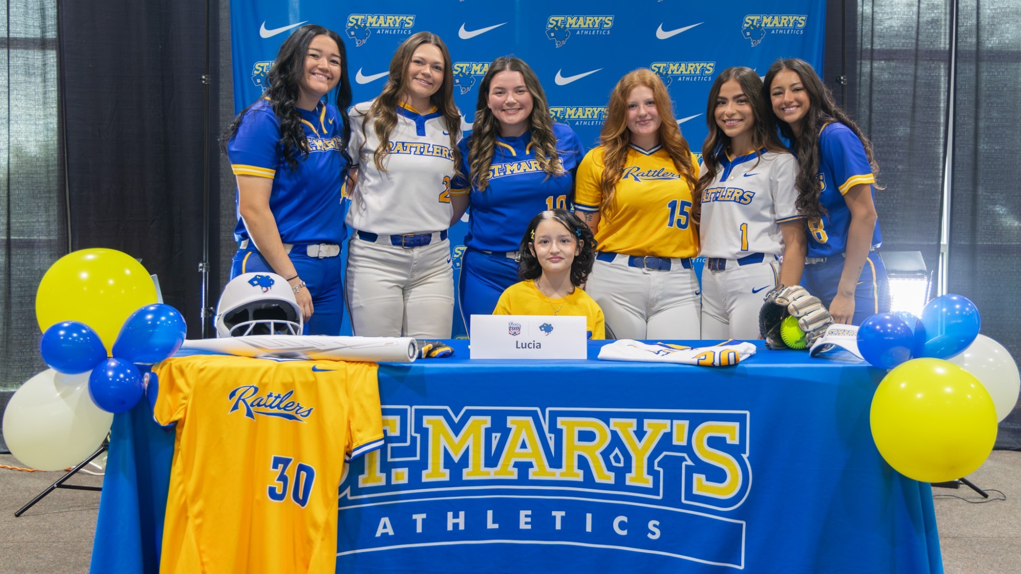 Six members of the softball team stand behind Lucia's chair on her signing day.