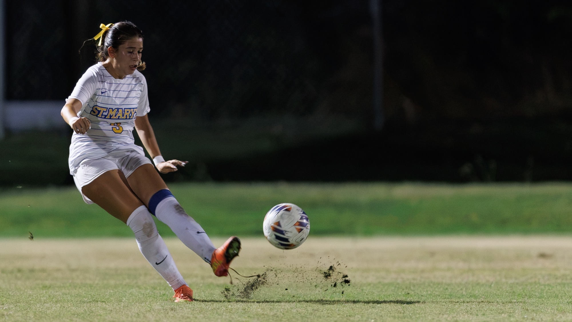 Alyssa Alba kicks the ball with her left foot as grass flies up with the ball.