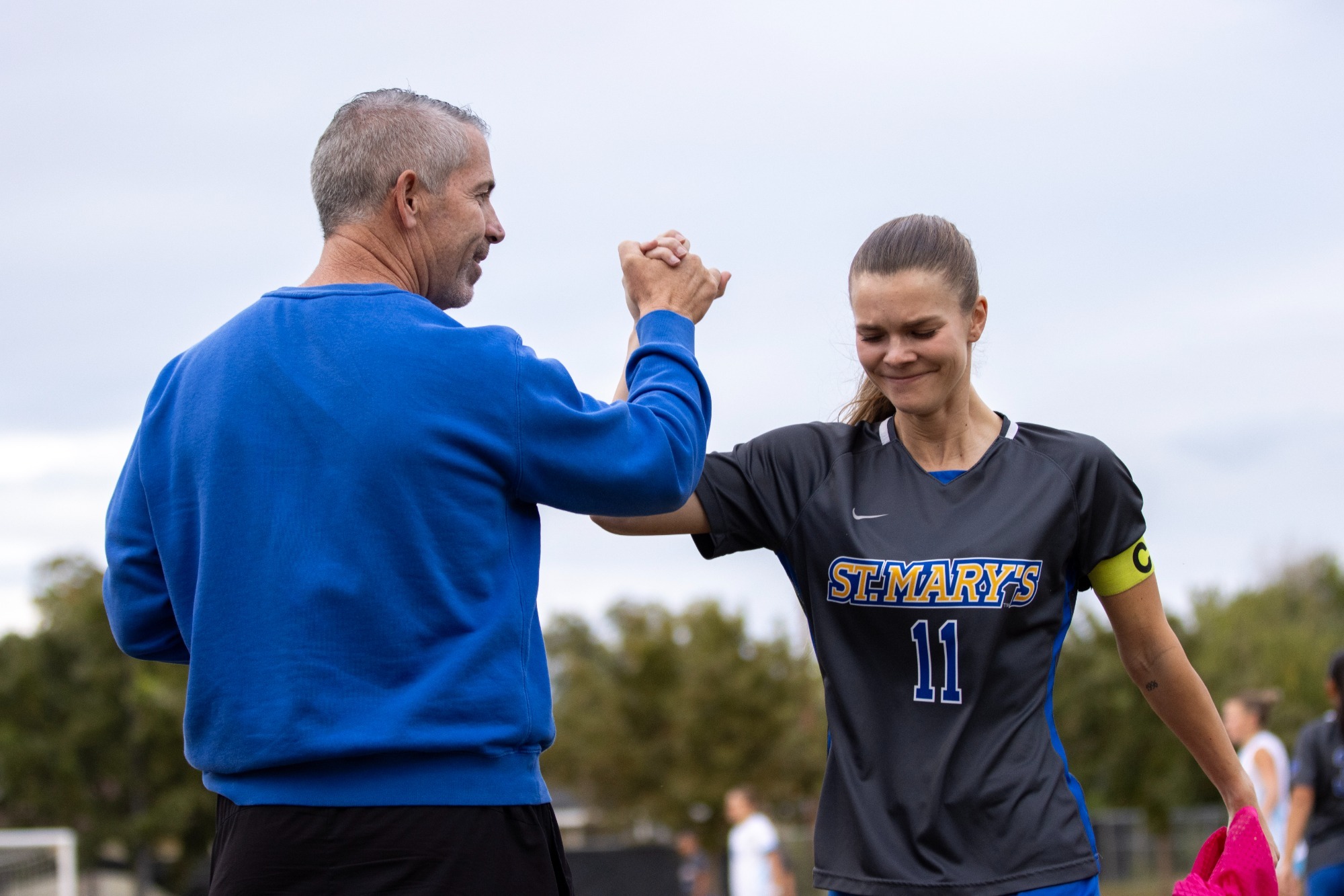 Head Coach Kevin Driggs and Elena Kossler clasp hands at shoulder level as Kossler leaves the pitch for the final time in her career.