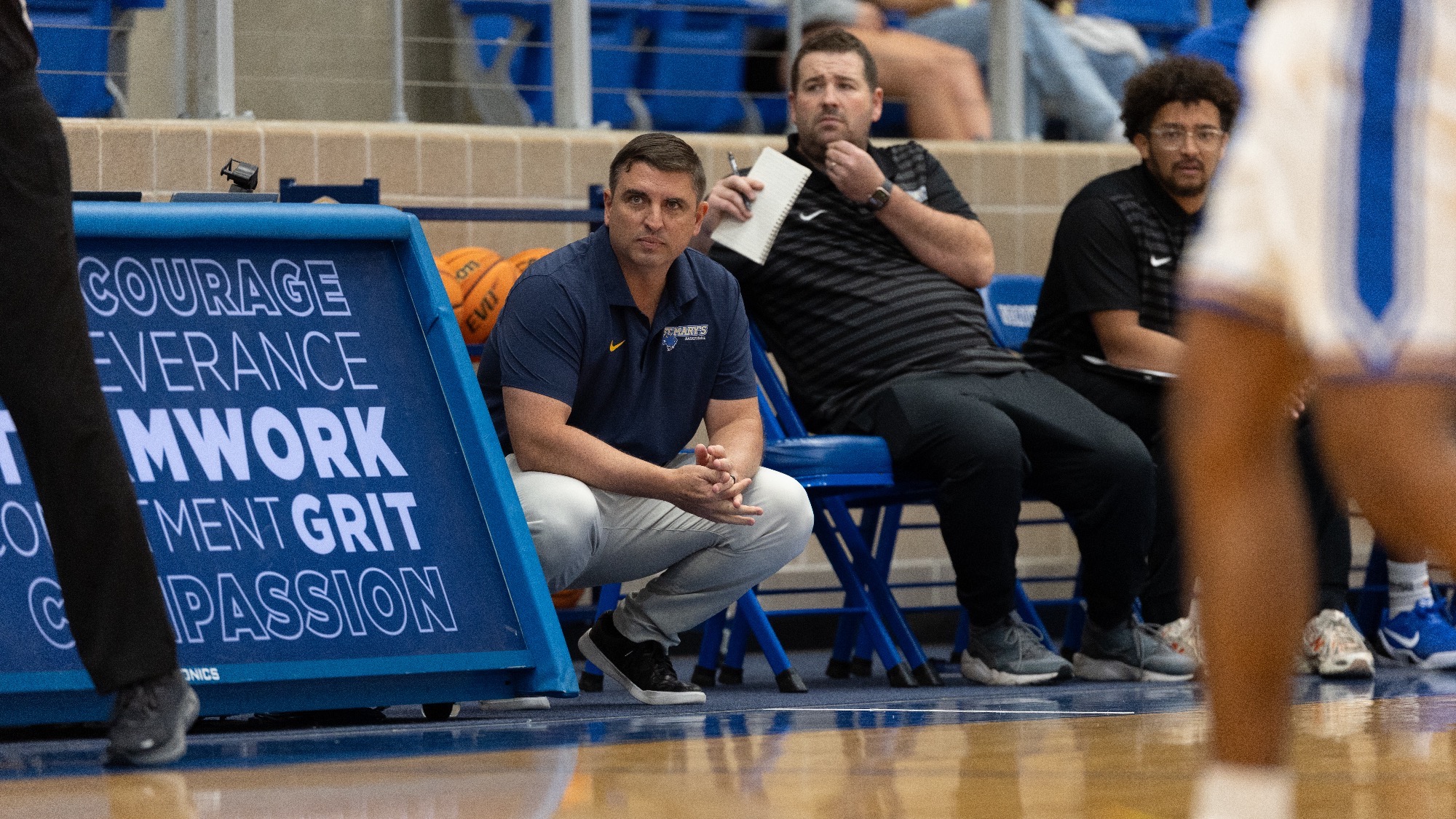 Head Coach Bubba Meyer crouches in front of the bench, with his assistant coaches sitting off his right shoulder.