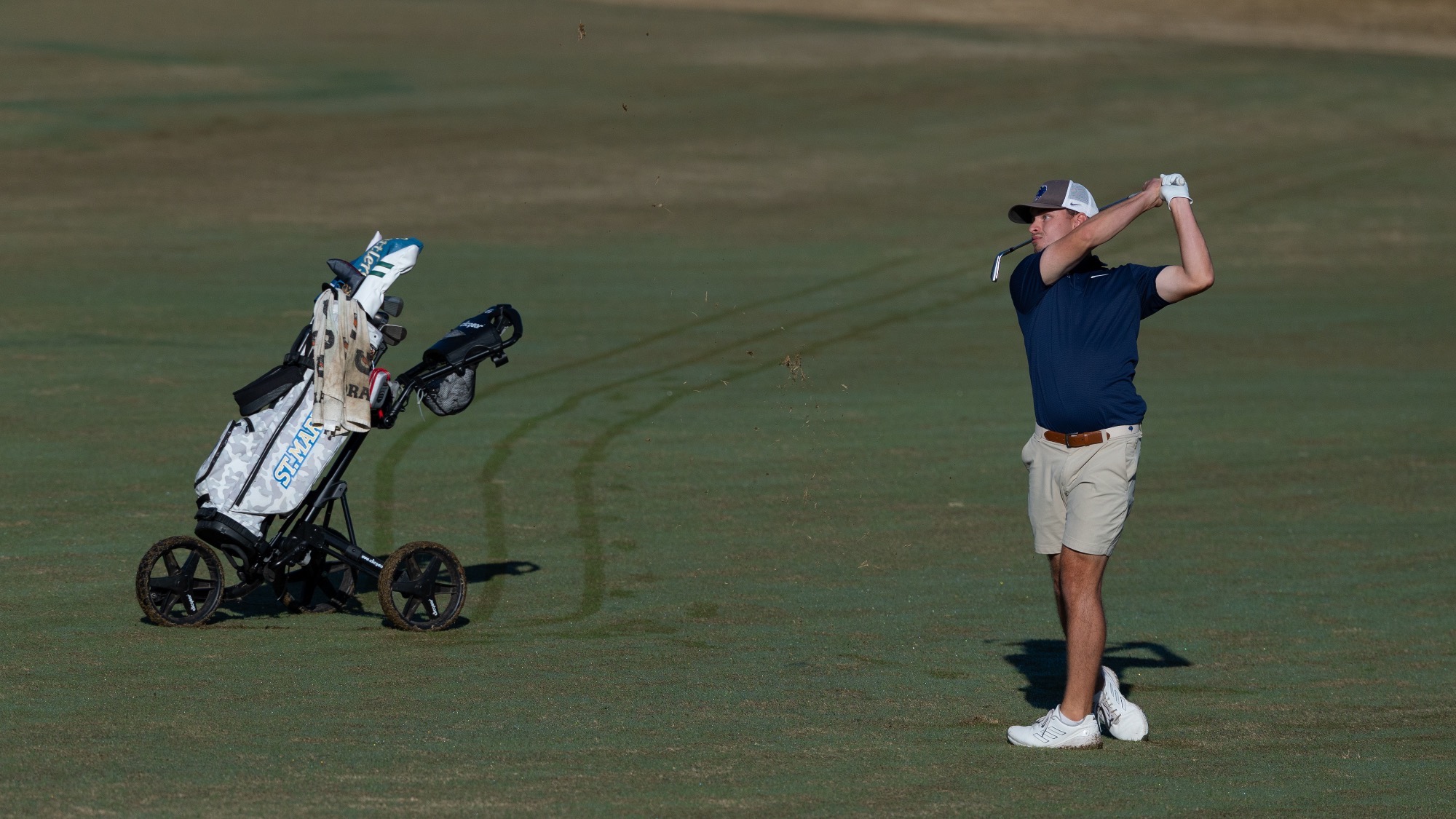 Connor Smith hits a shot from the fairway with his bag off to the side and the track marks from his cart in the grass.