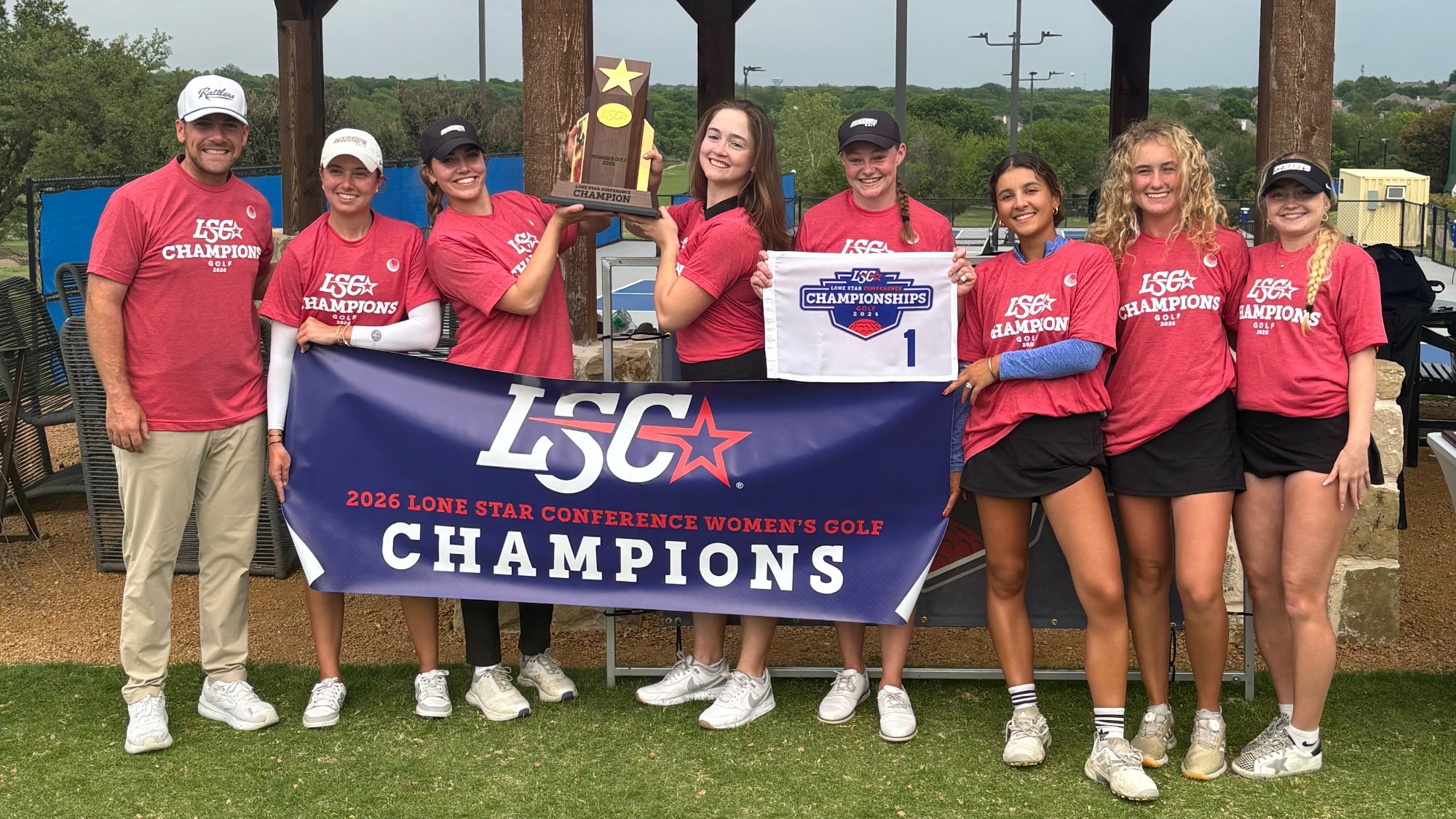 The 2026 Women's Golf team stands in a line with the trophy and the LSC Champions banner in front of them.
