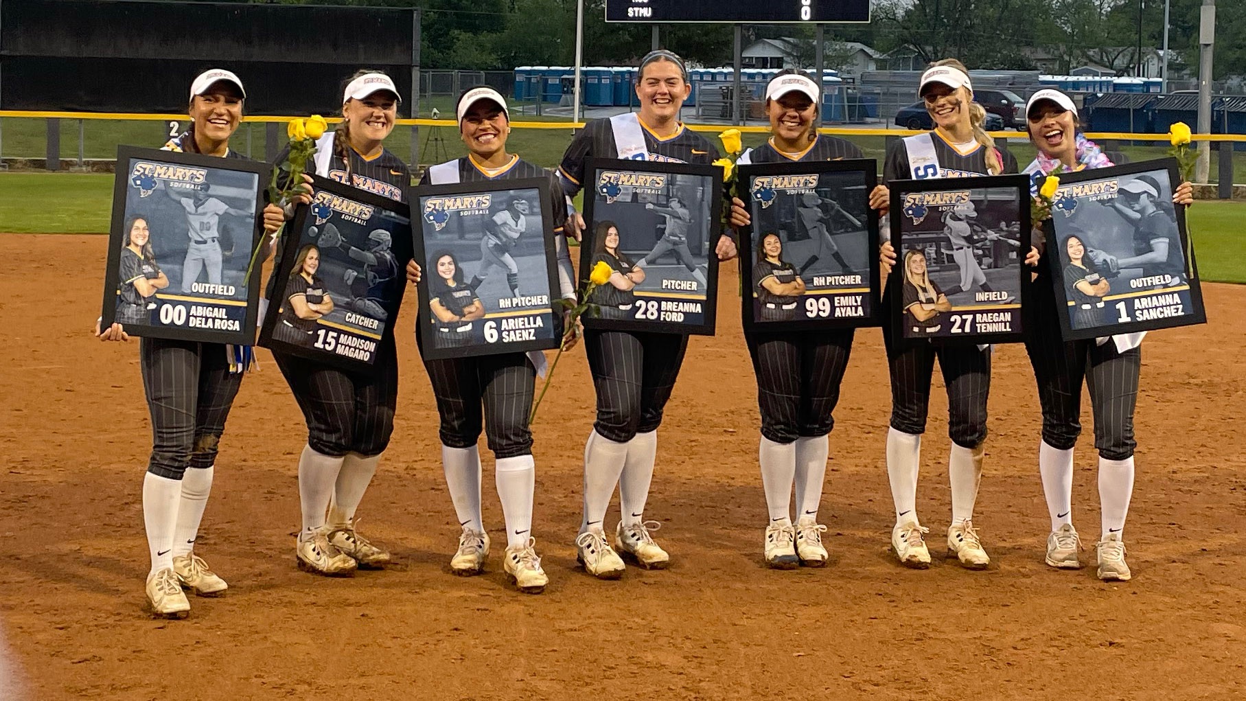 The seven seniors stand in the pitching circle with their senior posters and a yellow rose.