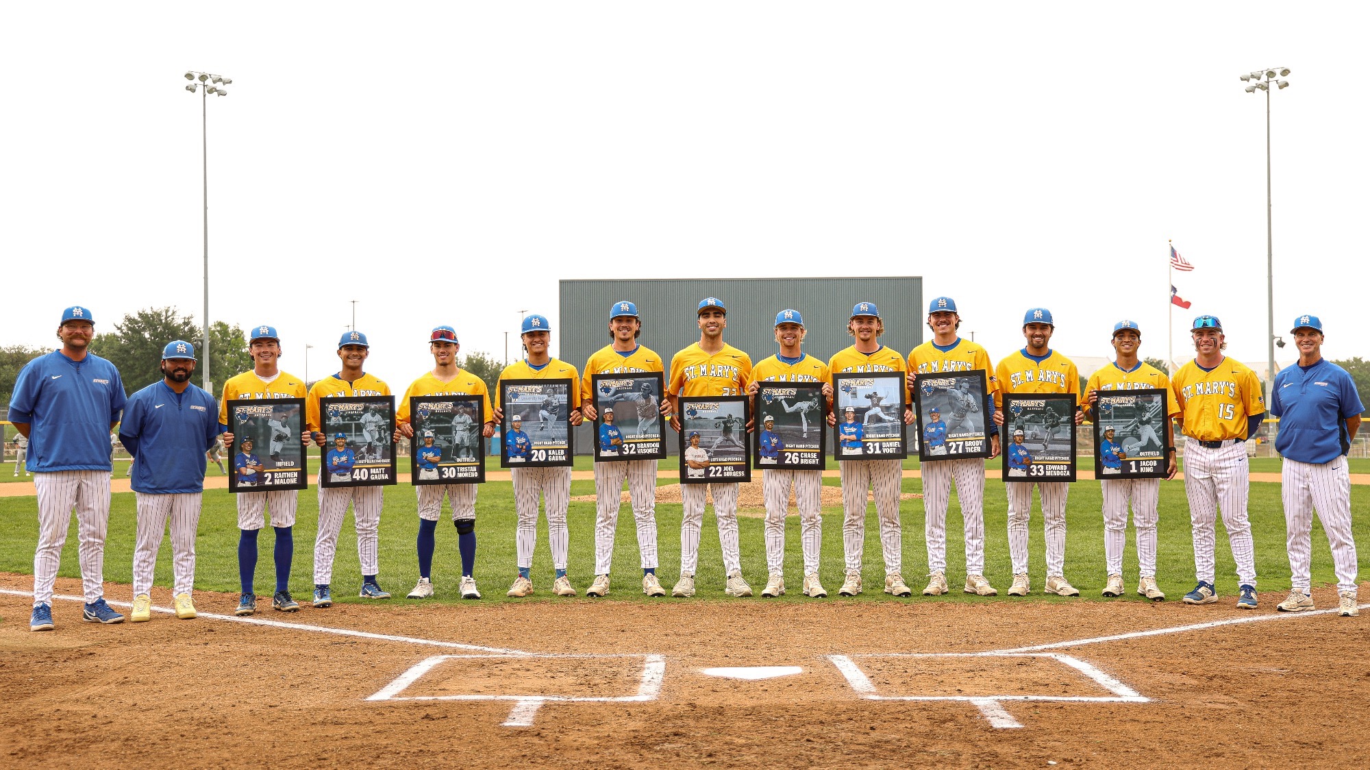 The coaching staff and seniors stand between home plate and the pitching mound with their senior posters.