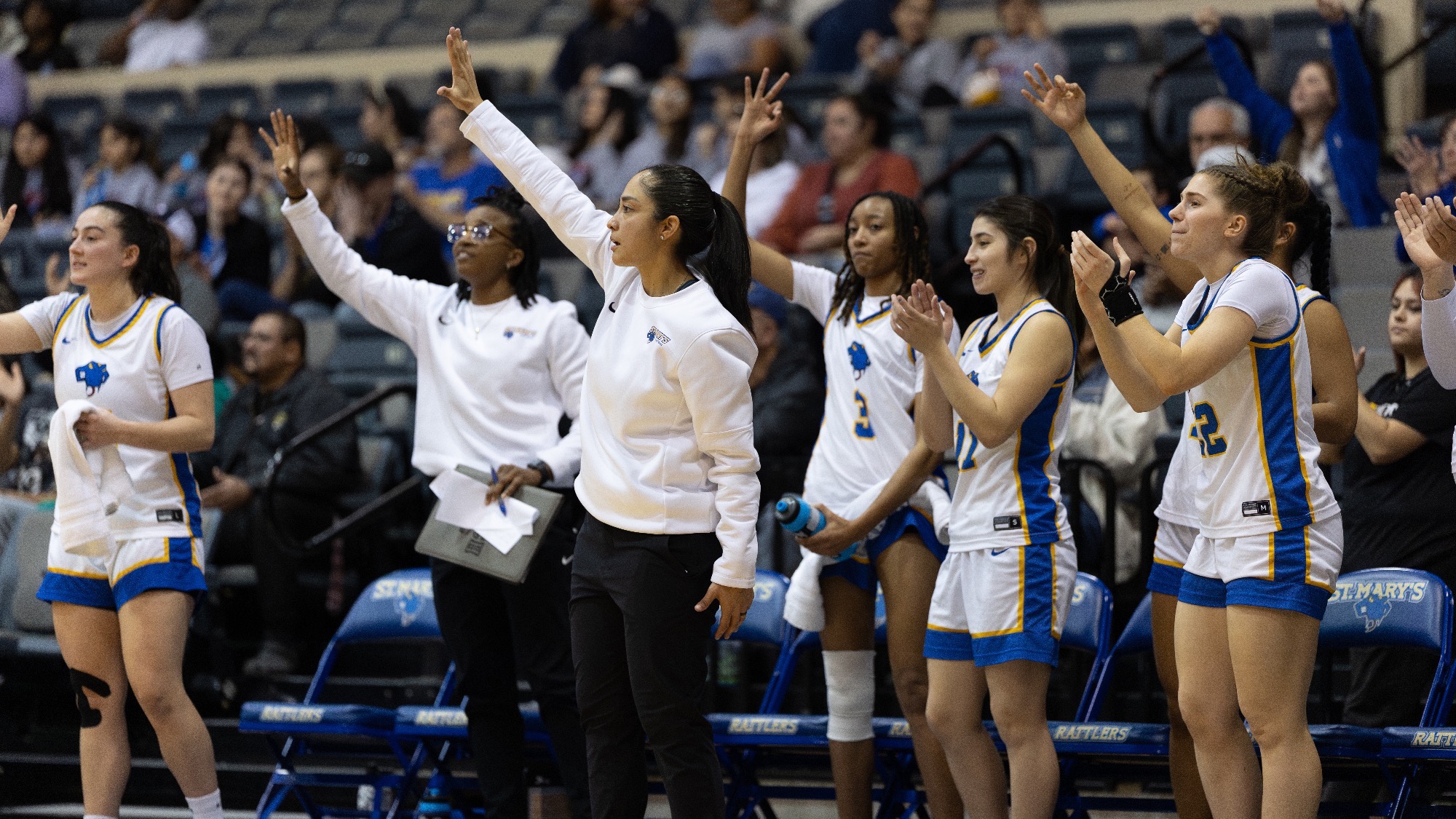 Head Coach Val Huizar stands in front of the bench calling a play, while the players celebrate a three-pointer.
