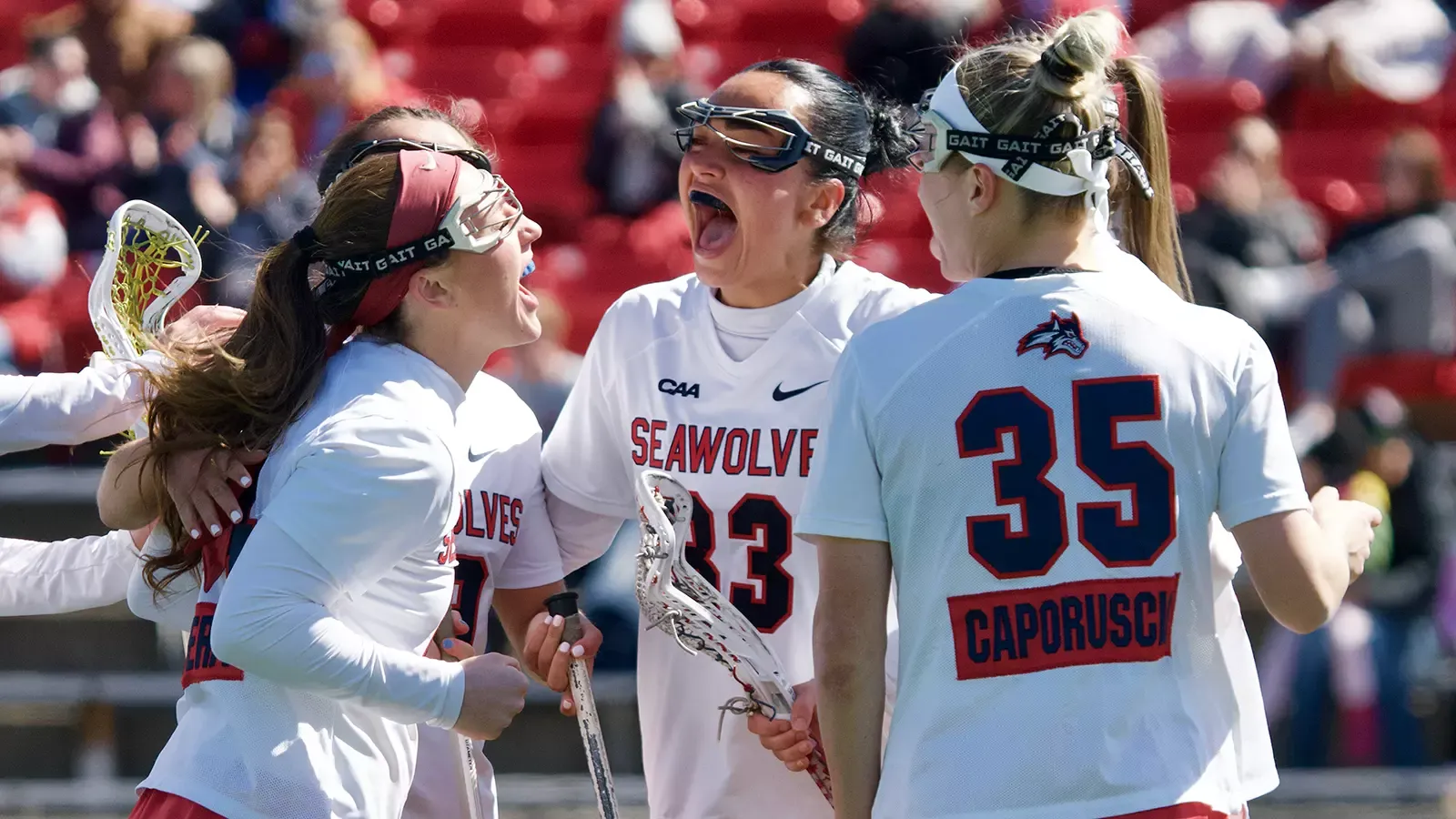 Stony Brook women's lacrosse players celebrating a goal. Mirabella Altebrando is forward facing in a huddle celebrating.