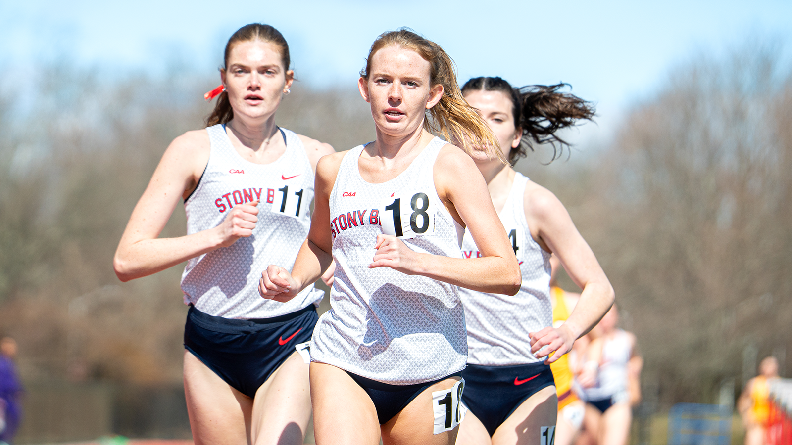 Grace Weigele running on the track in white red and blue Stony Brook uniform with two other runners trailing behind her. 