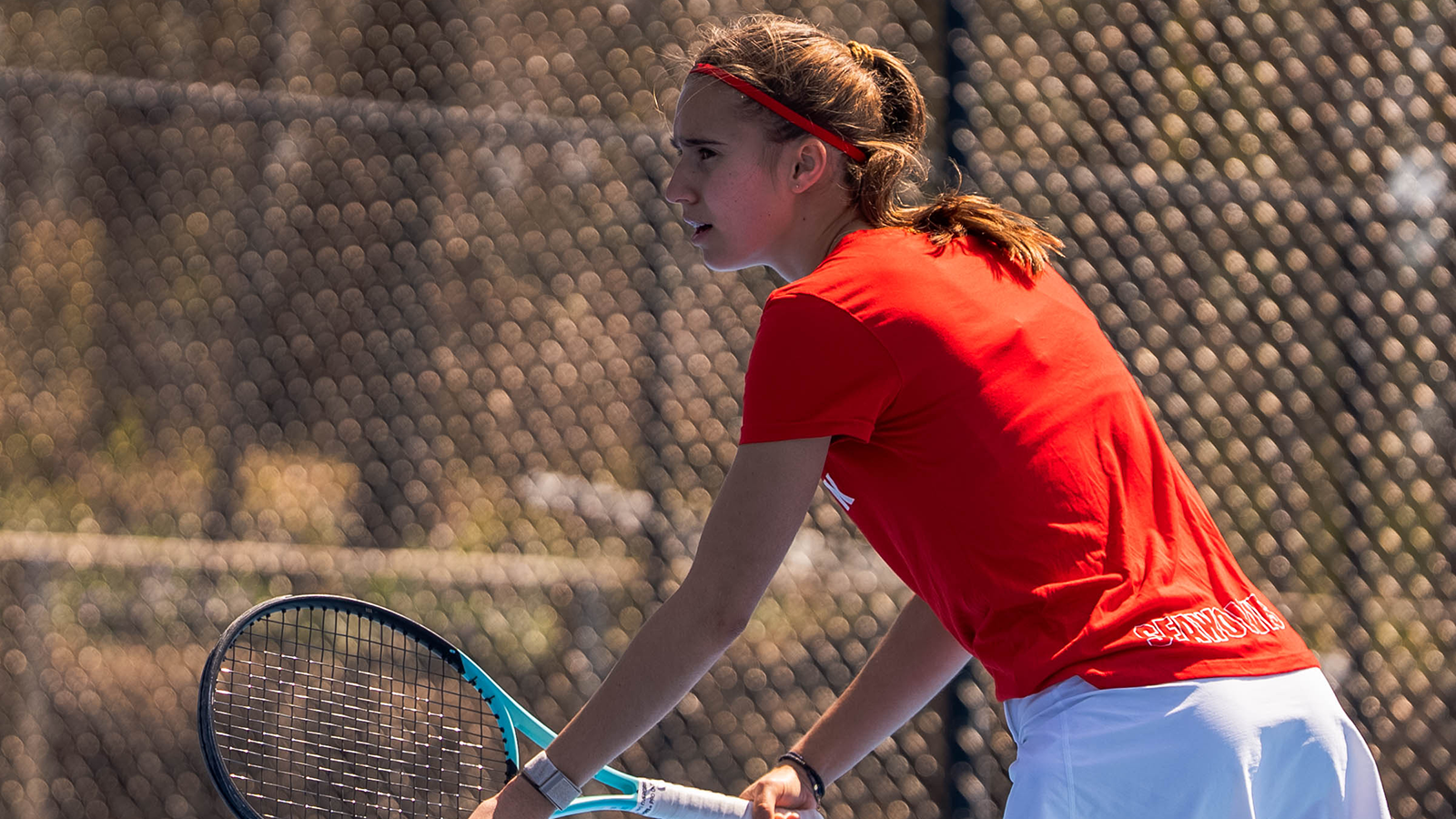 Mila Charles in a red Stony Brook tennis uniform preparing to throw the ball up and start her serve attempt in a match. 