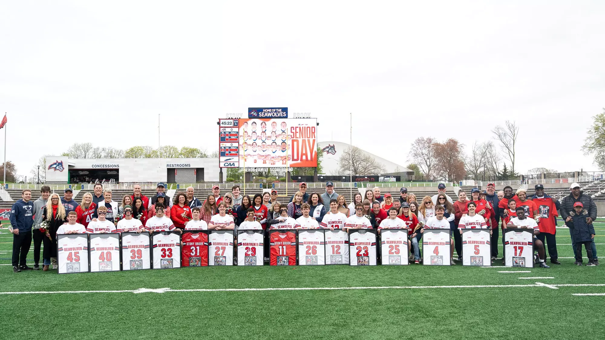Group photo of the 2026 Stony Brook men's lacrosse senior class on the LaValle Stadium turf with their families. The players hold framed jerseys and in the backdrop is the videoboard with a graphic of the senior class.