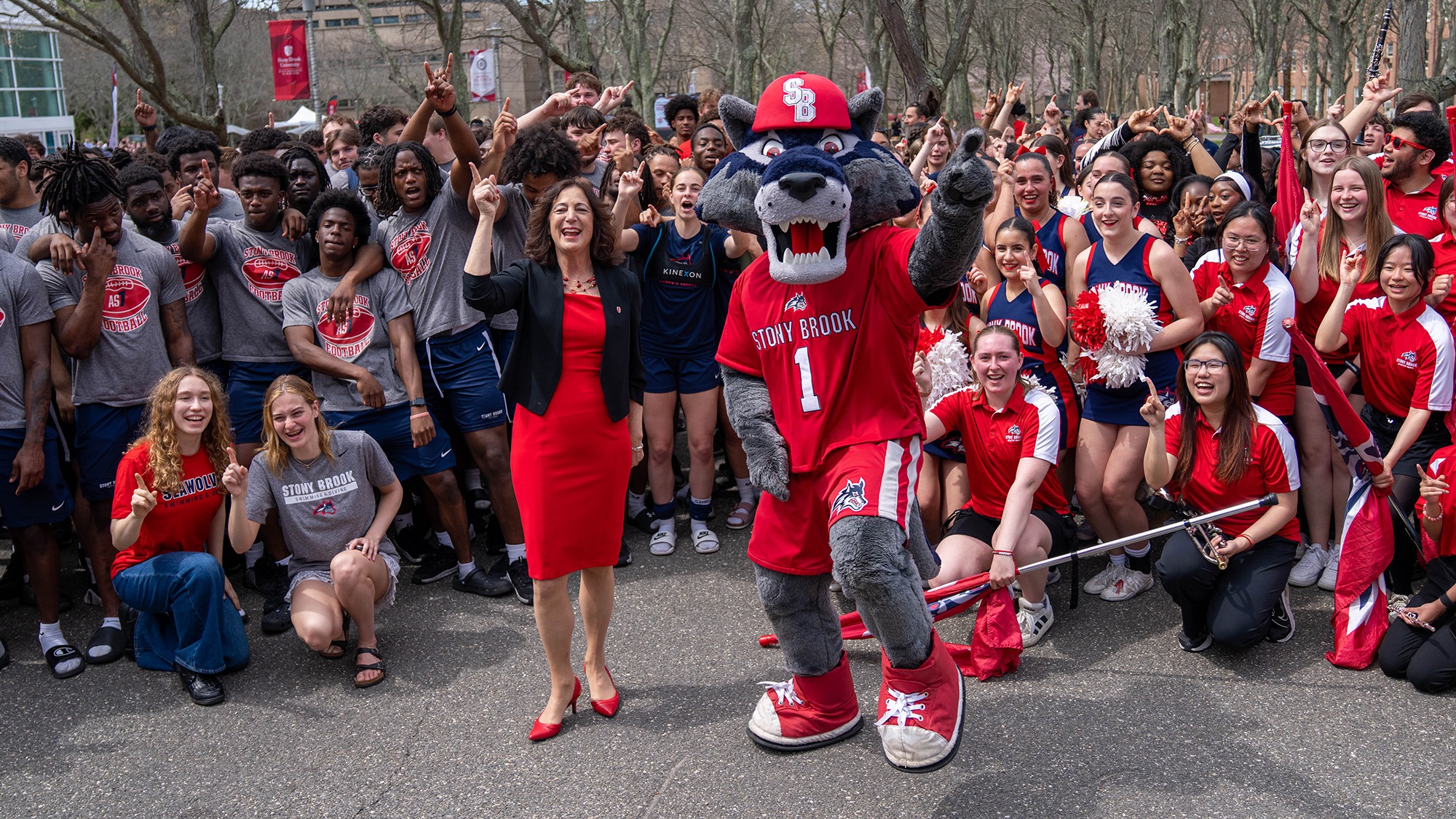 Stony Brook University President Andrea Goldsmith and mascot Wolfie, flanked by a large group of Stony Brook student-athletes and members of the spirit squad at an outdoor location on campus.