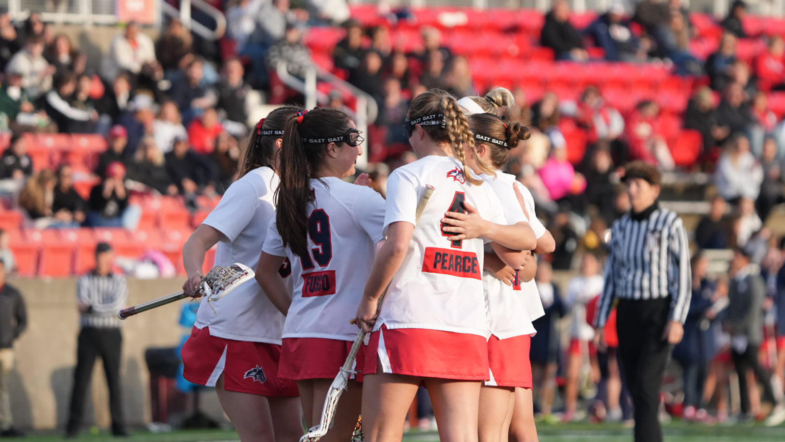 Stony Brook women's lacrosse players Julia Fusco and Emma Pearce huddle around their teammates to celebrate a goal. 