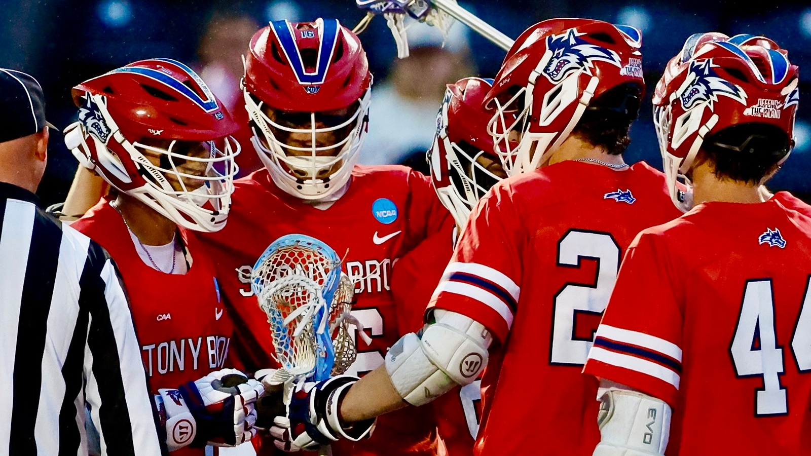 Stony Brook men's lacrosse players in red jersey's huddle around each other with the rain coming down around them. 