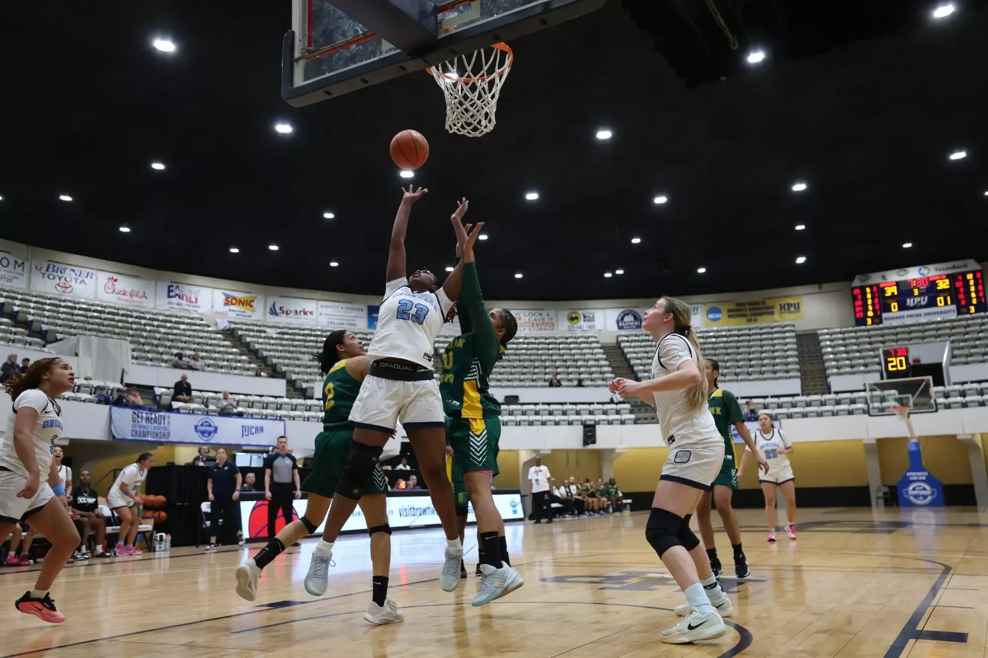Basketball game in action; player in white attempts a layup against green-clad defenders.