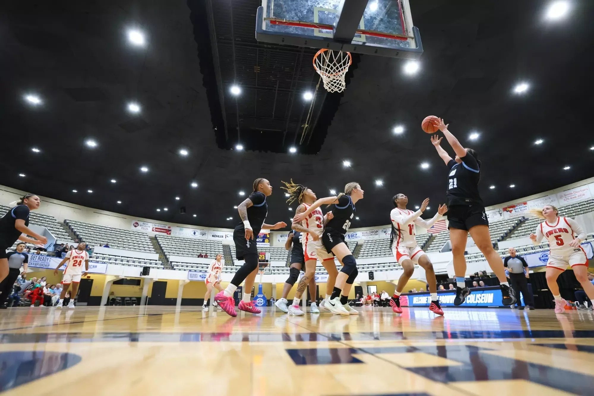 A women’s basketball player in a black uniform jumps to shoot the ball near the hoop, surrounded by players in white uniforms. 
