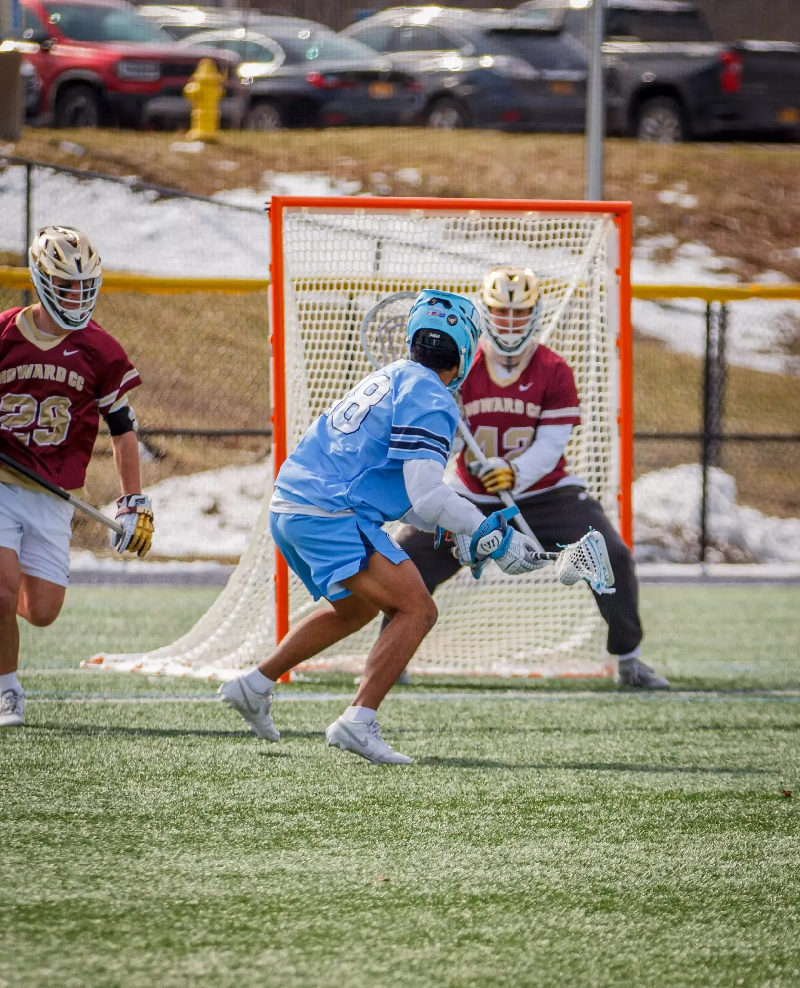 Lacrosse players in action: a player in a blue uniform attempts a shot at the goal, defended by a goalie in maroon.