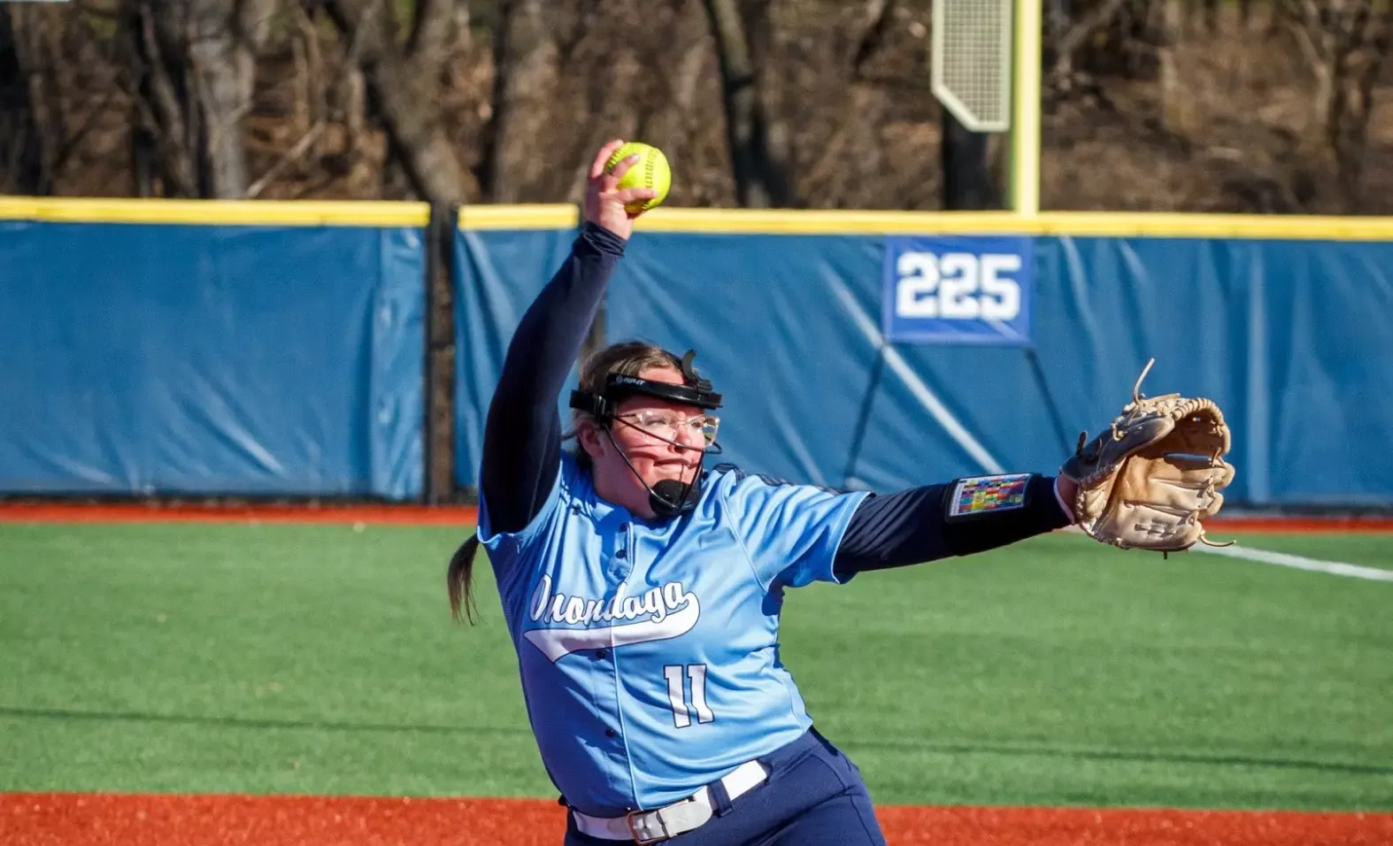 Softball player in a light blue jersey winds up to pitch on a sunny day. 