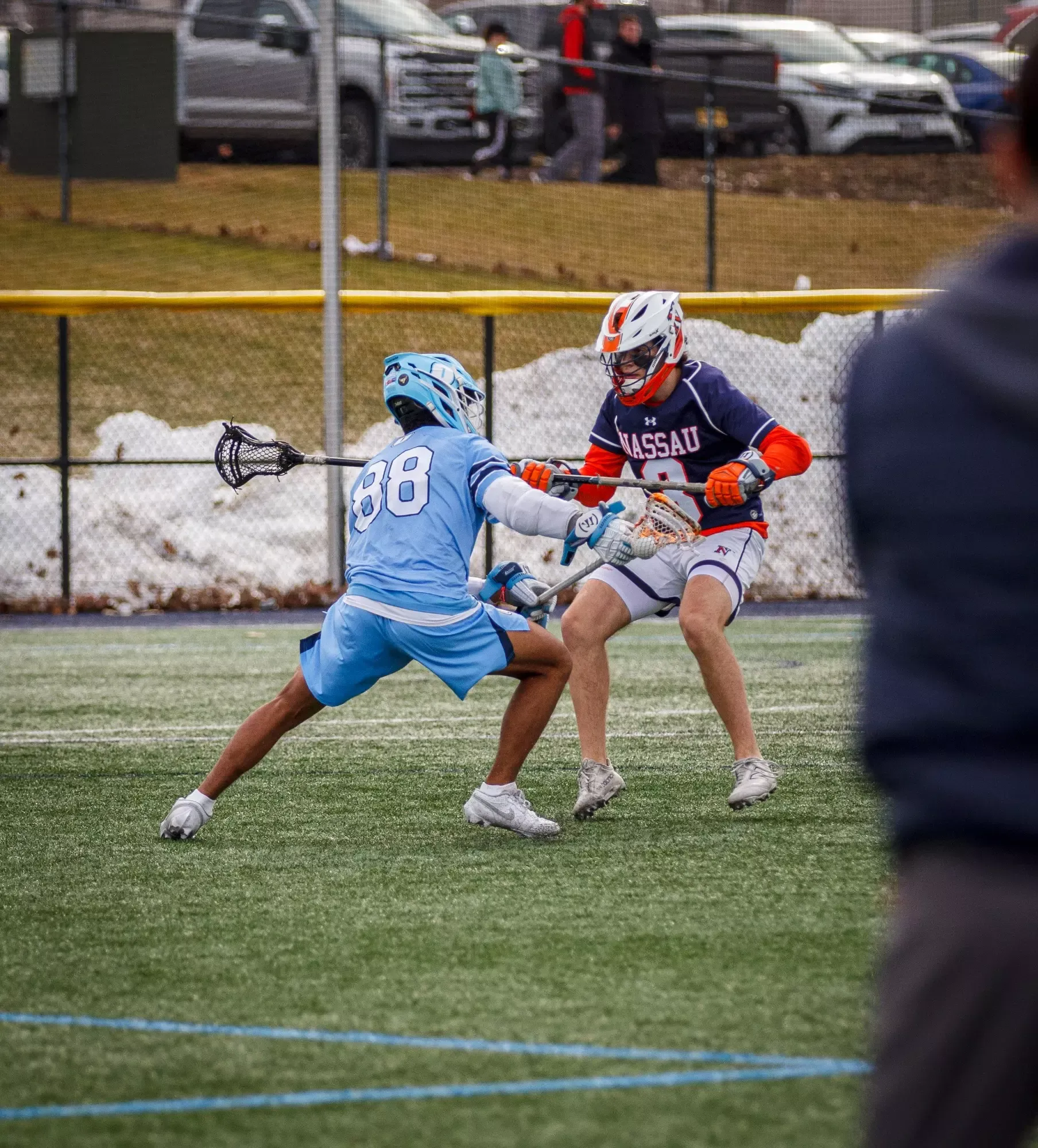 Two lacrosse players in action on a field. One in light blue attempts to block while the other in navy, labeled 