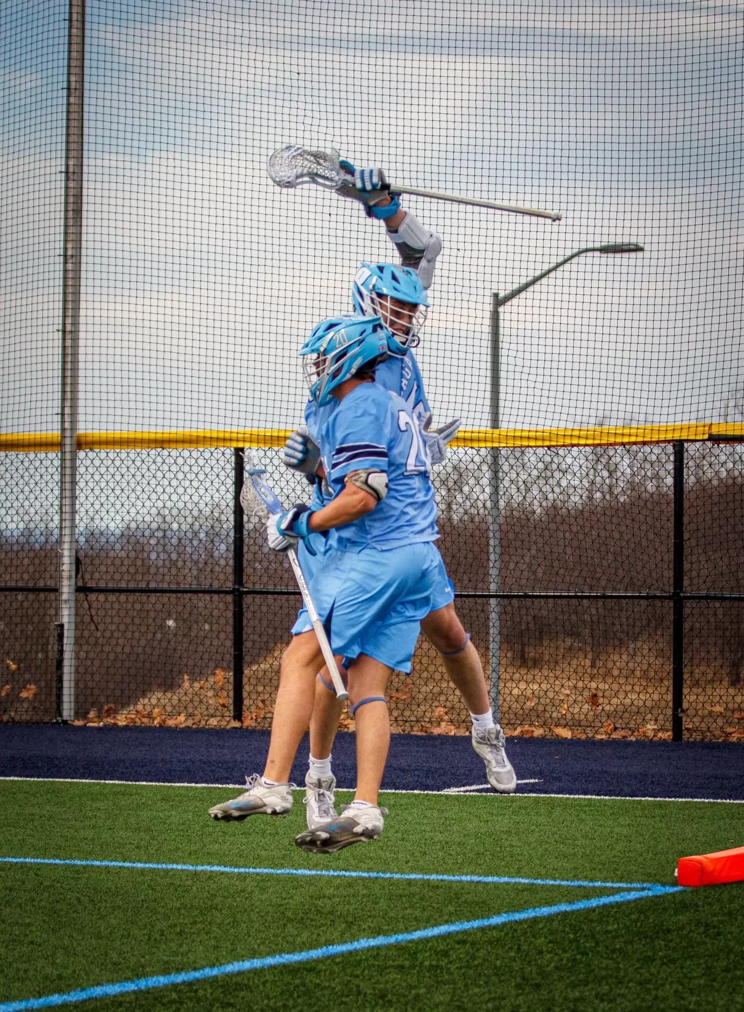 Two lacrosse players in light blue uniforms celebrate a scoring play on a green field.