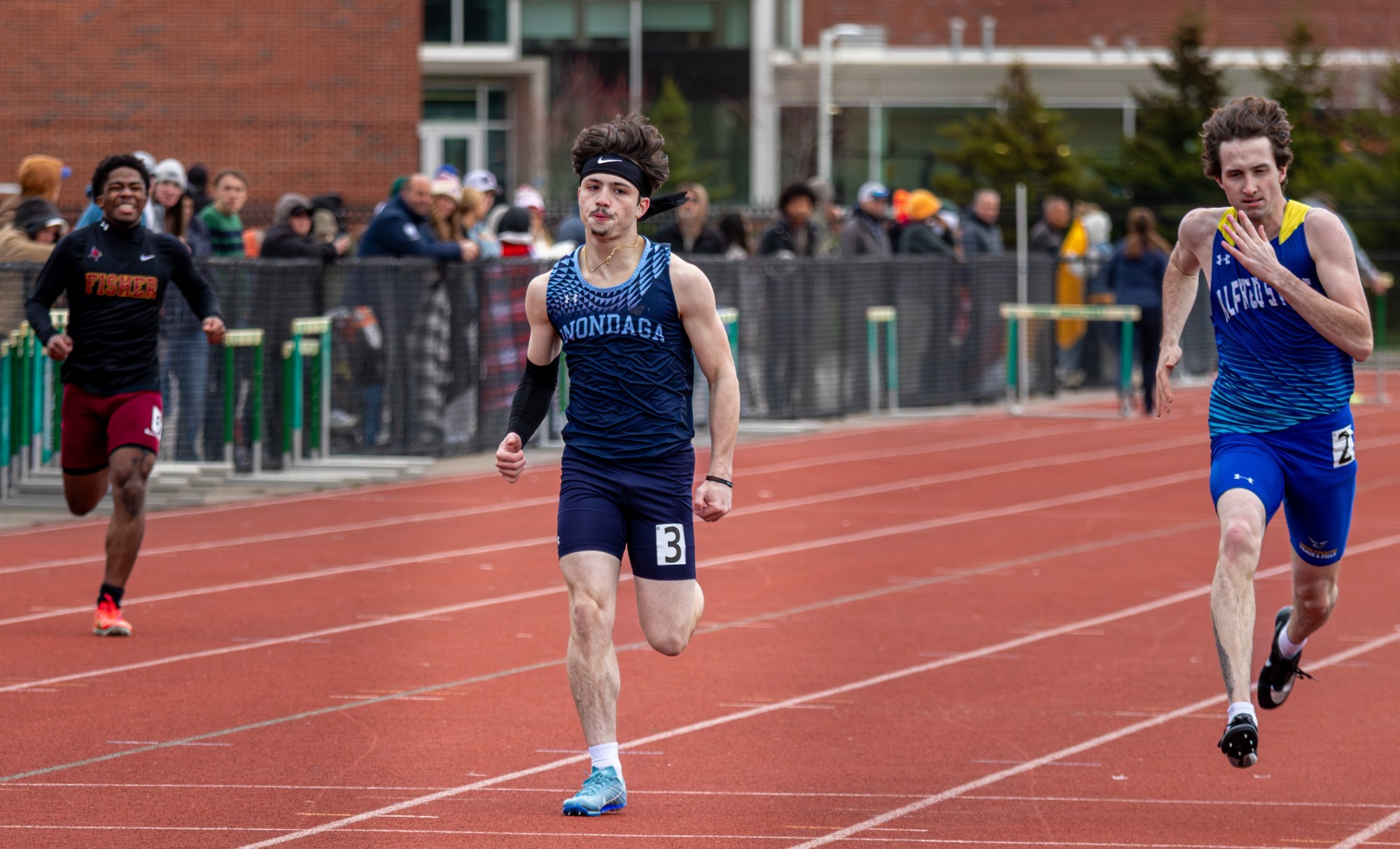 Three runners wear distinct uniforms, with a blurred crowd watching from the sideline under cloudy skies.