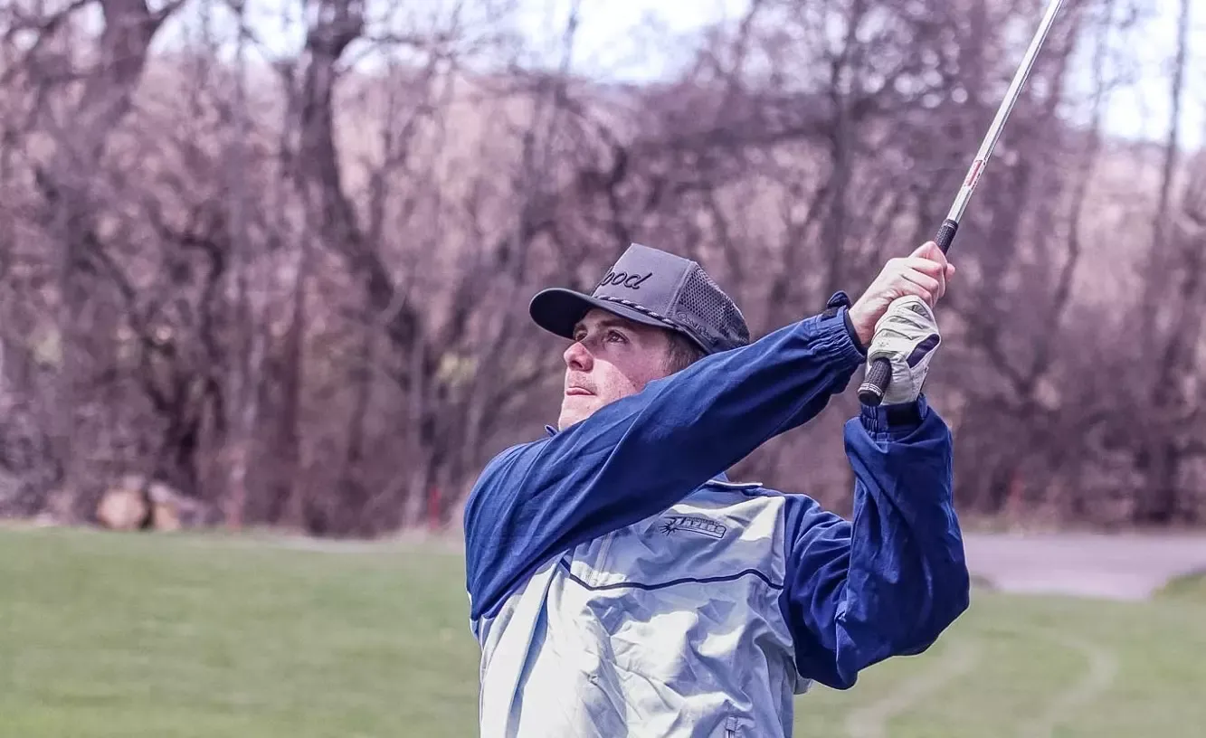 Golfer in mid-swing on a grassy course, wearing a cap and jacket, with a focused expression.