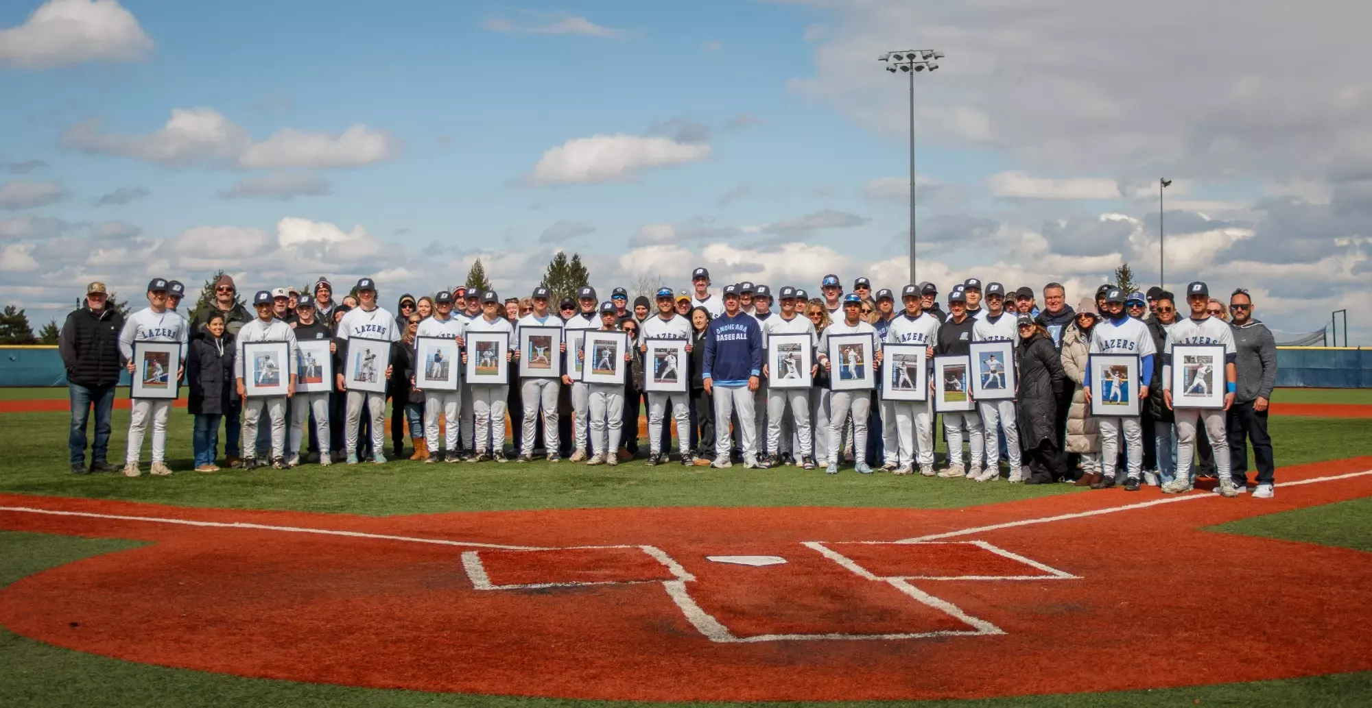 A baseball team poses on a field, holding framed photos. They're smiling under a partly cloudy sky, creating a celebratory and proud atmosphere.