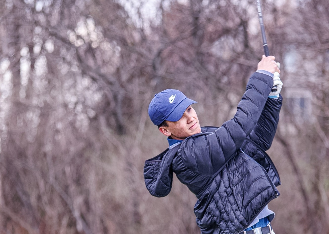 A golfer in a cap and jacket swings a club on a green field, surrounded by leafless trees.
