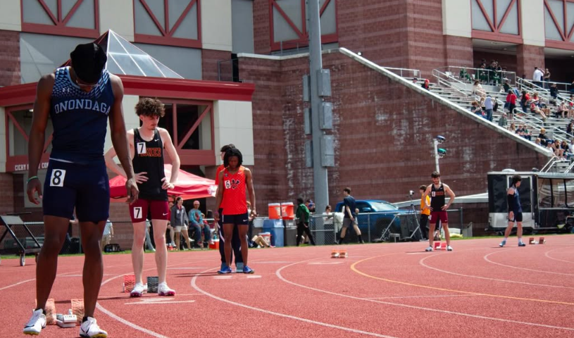 Athletes in track uniforms prepare at the starting line on a sunny day. 