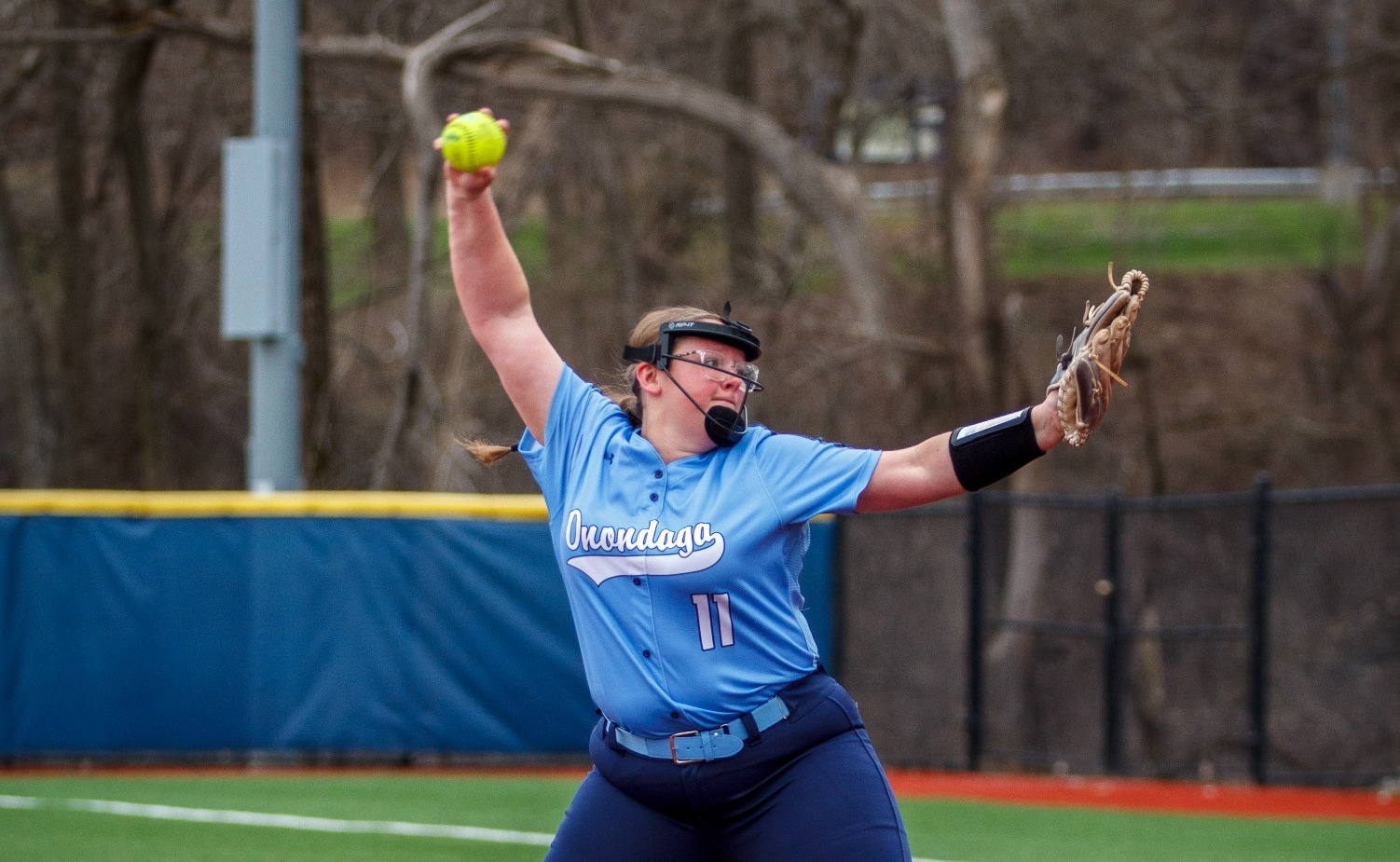 Softball player in blue uniform pitching on a red field with determination. Her arm is raised mid-throw, emphasizing focus and athleticism.