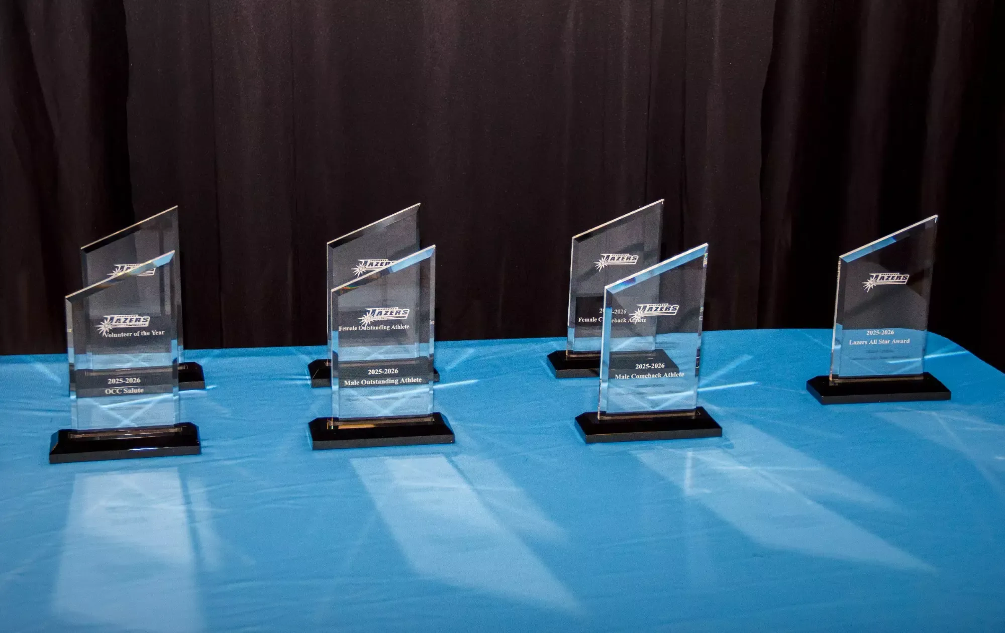 Clear glass awards with black bases are neatly arranged on a blue table, set against a black curtain backdrop, creating an elegant and celebratory atmosphere.