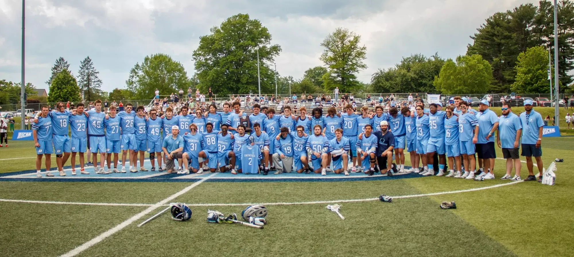 A large group of lacrosse players wearing light blue uniforms pose together on a field, smiling. Lacrosse sticks and helmets lie on the grass.