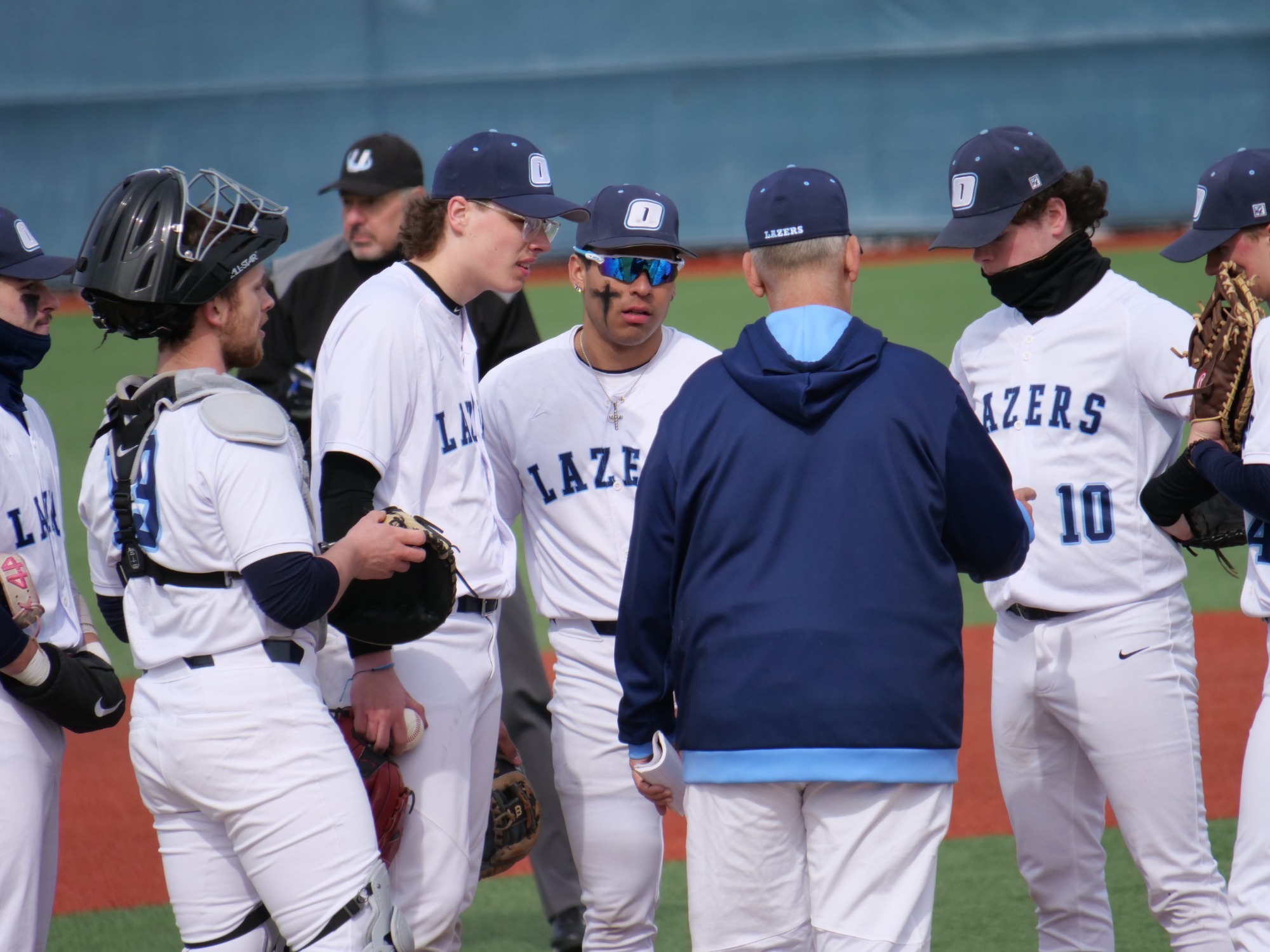 A baseball team huddles on a field, players in white uniforms and blue caps, a coach in a blue jacket gives a pep talk. The mood is focused and determined.