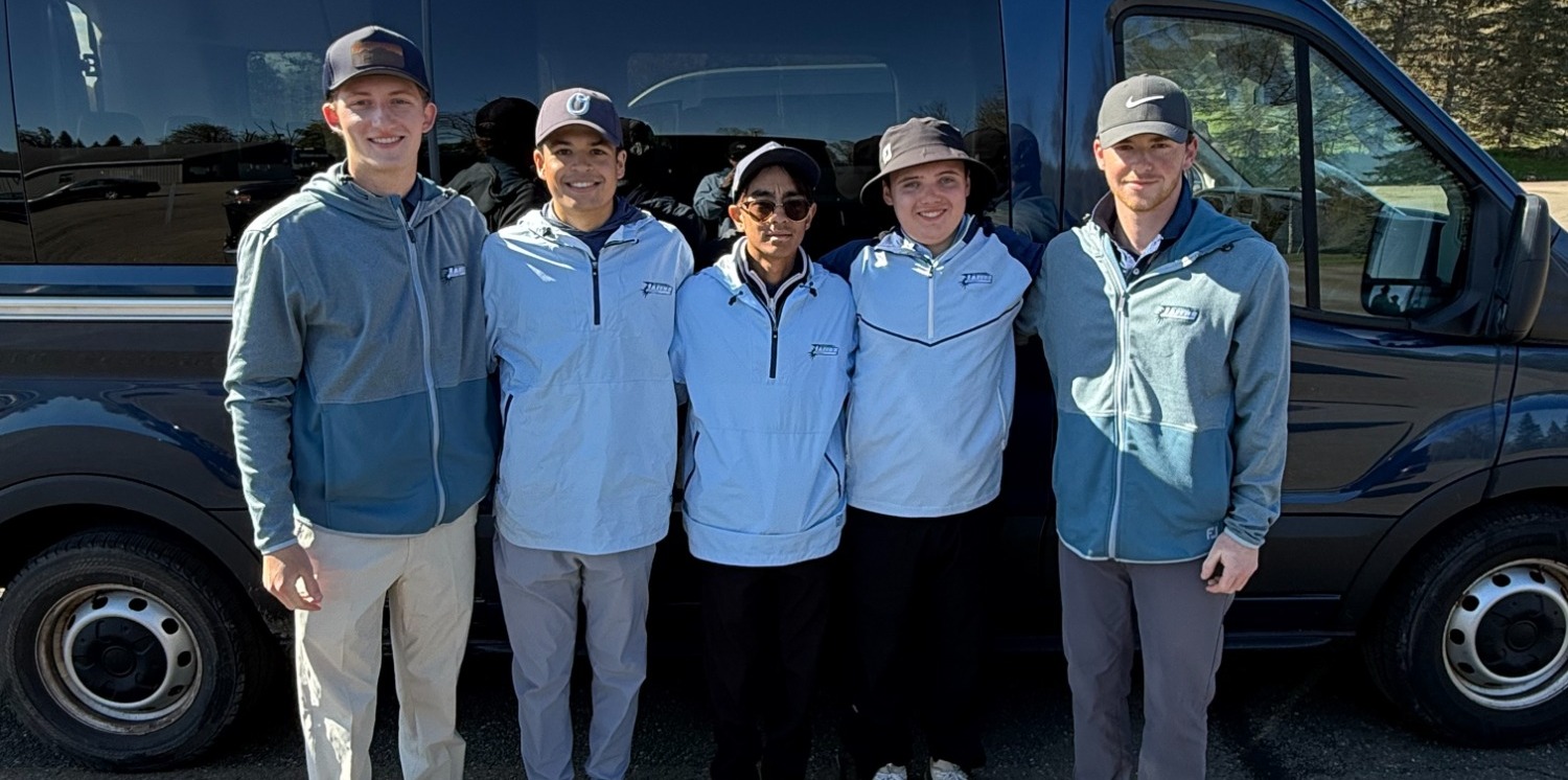 Five people stand smiling in front of a dark van on a sunny day. They wear matching light blue jackets and caps, conveying team spirit and camaraderie.