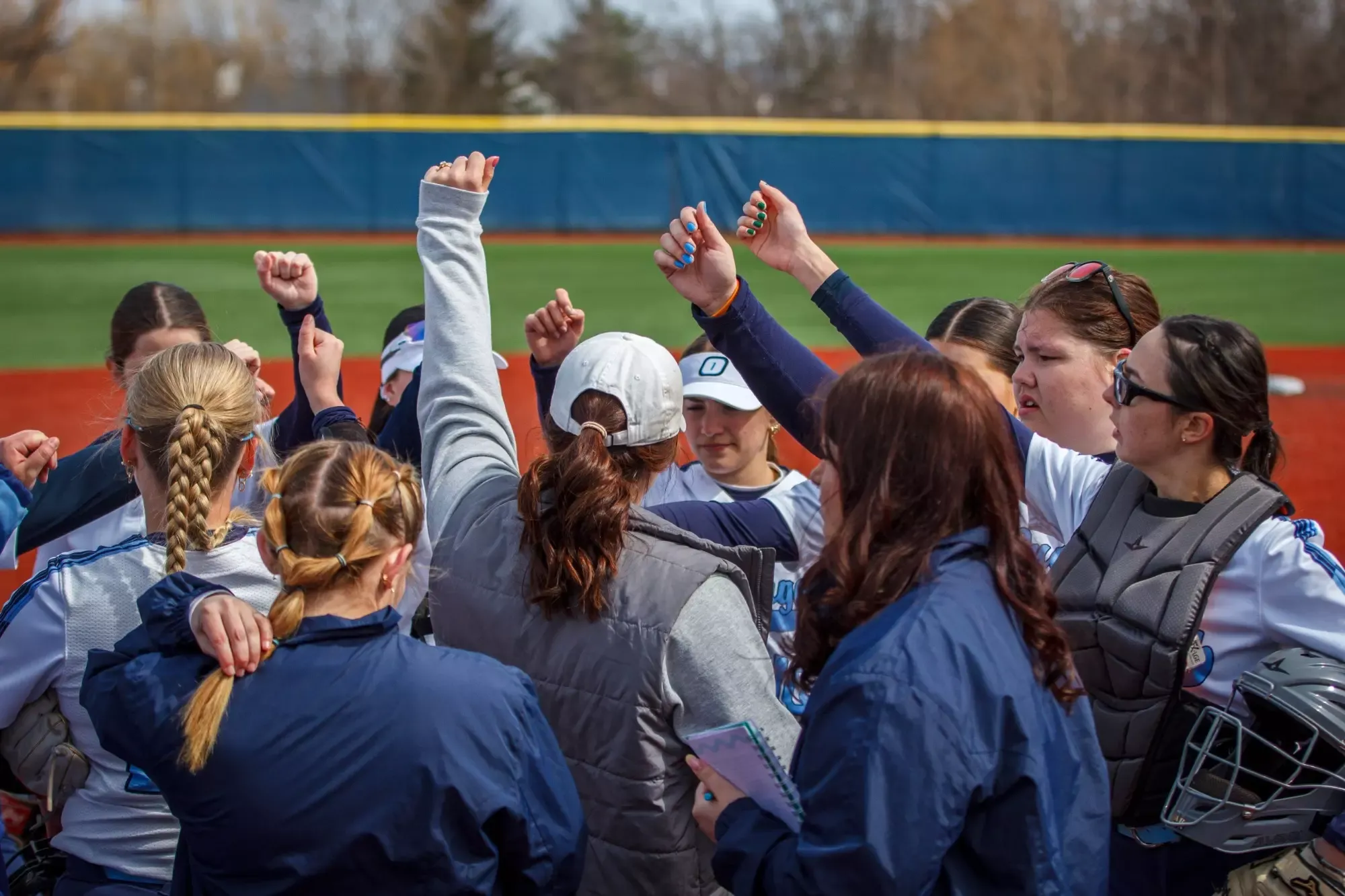 A softball team huddles together on a field, raising their fists in a show of unity and motivation. They wear uniforms and appear focused and spirited.