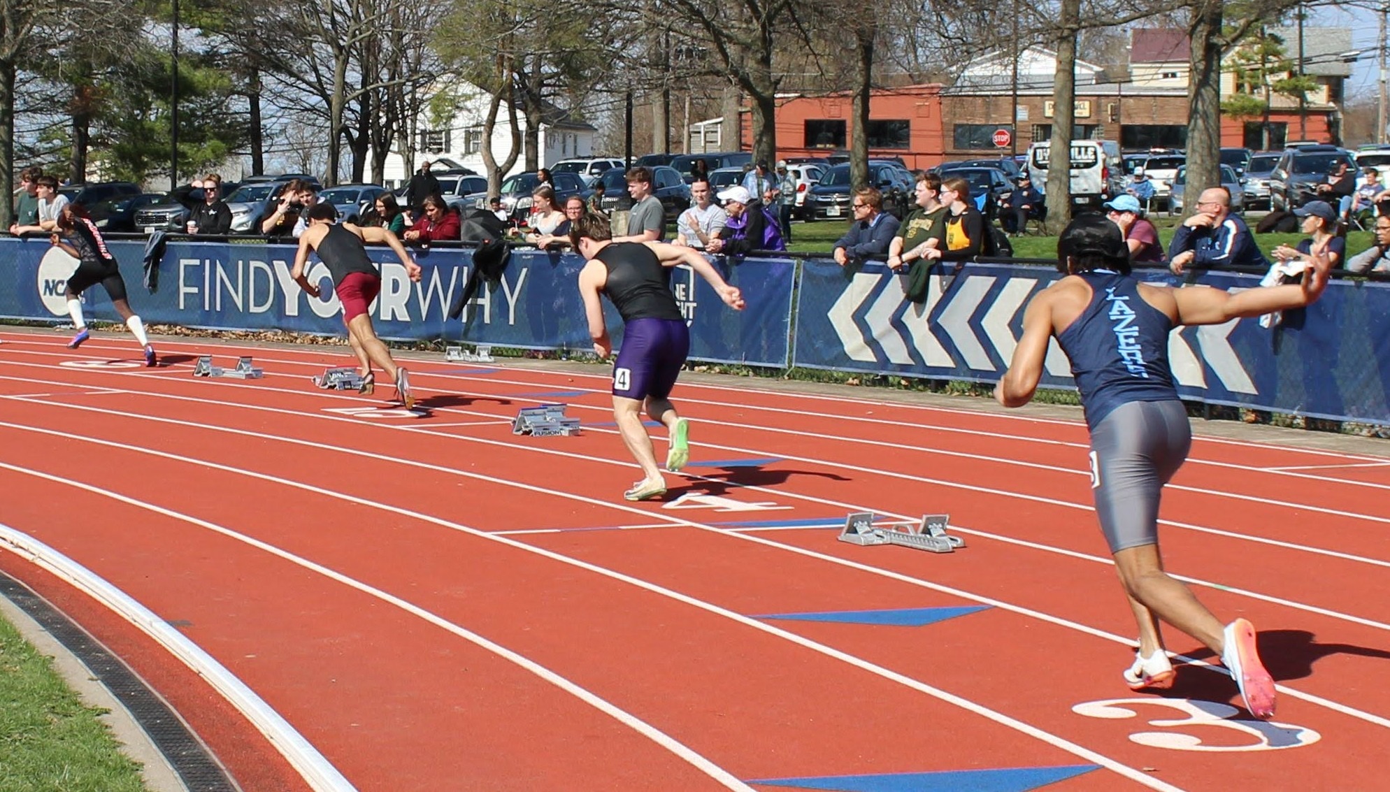 Runners sprint from the starting blocks on a sunlit outdoor track. Spectators watch from the sidelines, capturing the energy and competitive spirit.