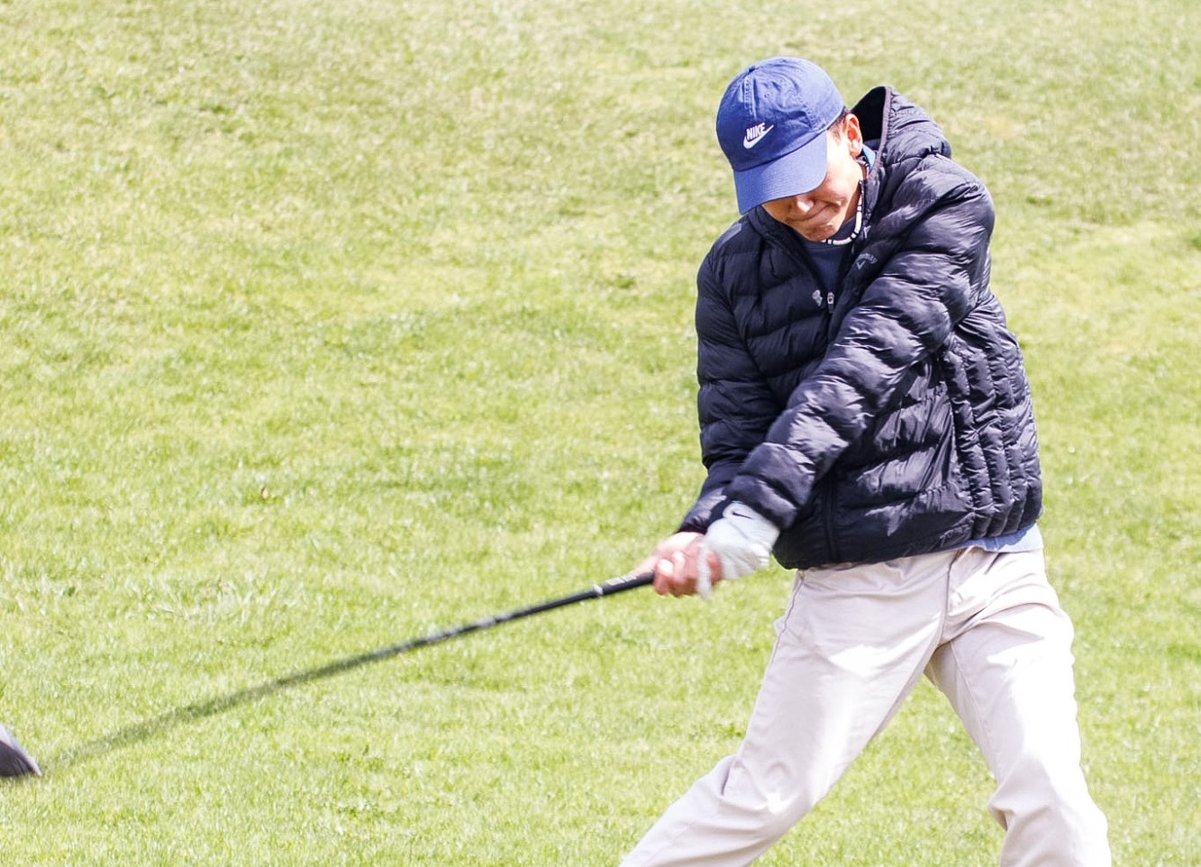 A golfer in a black jacket and blue cap swings a club on a sunny day, surrounded by lush green grass. The scene conveys focus and concentration.