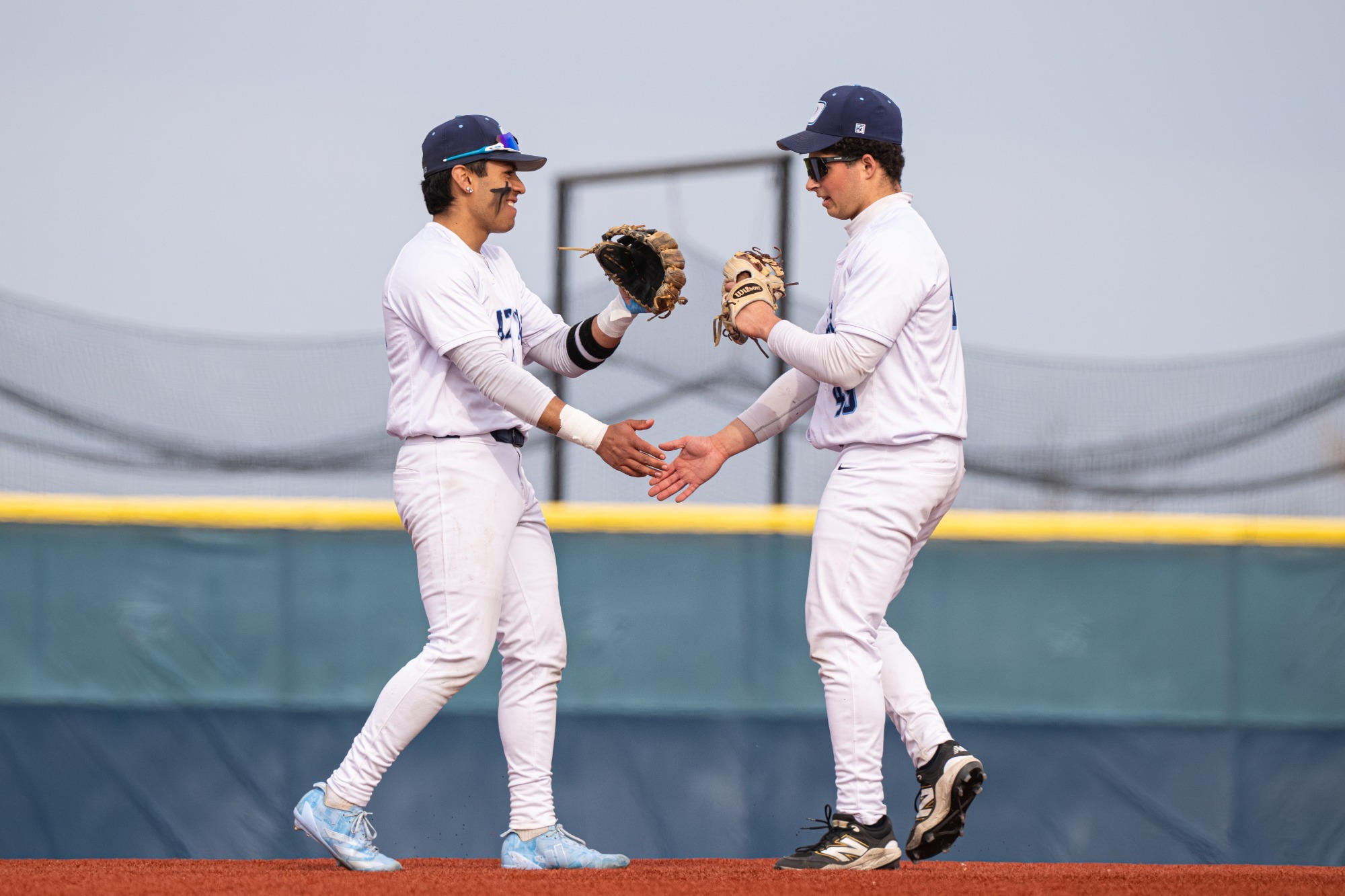 Two baseball players in white uniforms exchange a handshake on a field, smiling. Their camaraderie suggests teamwork and a positive game.