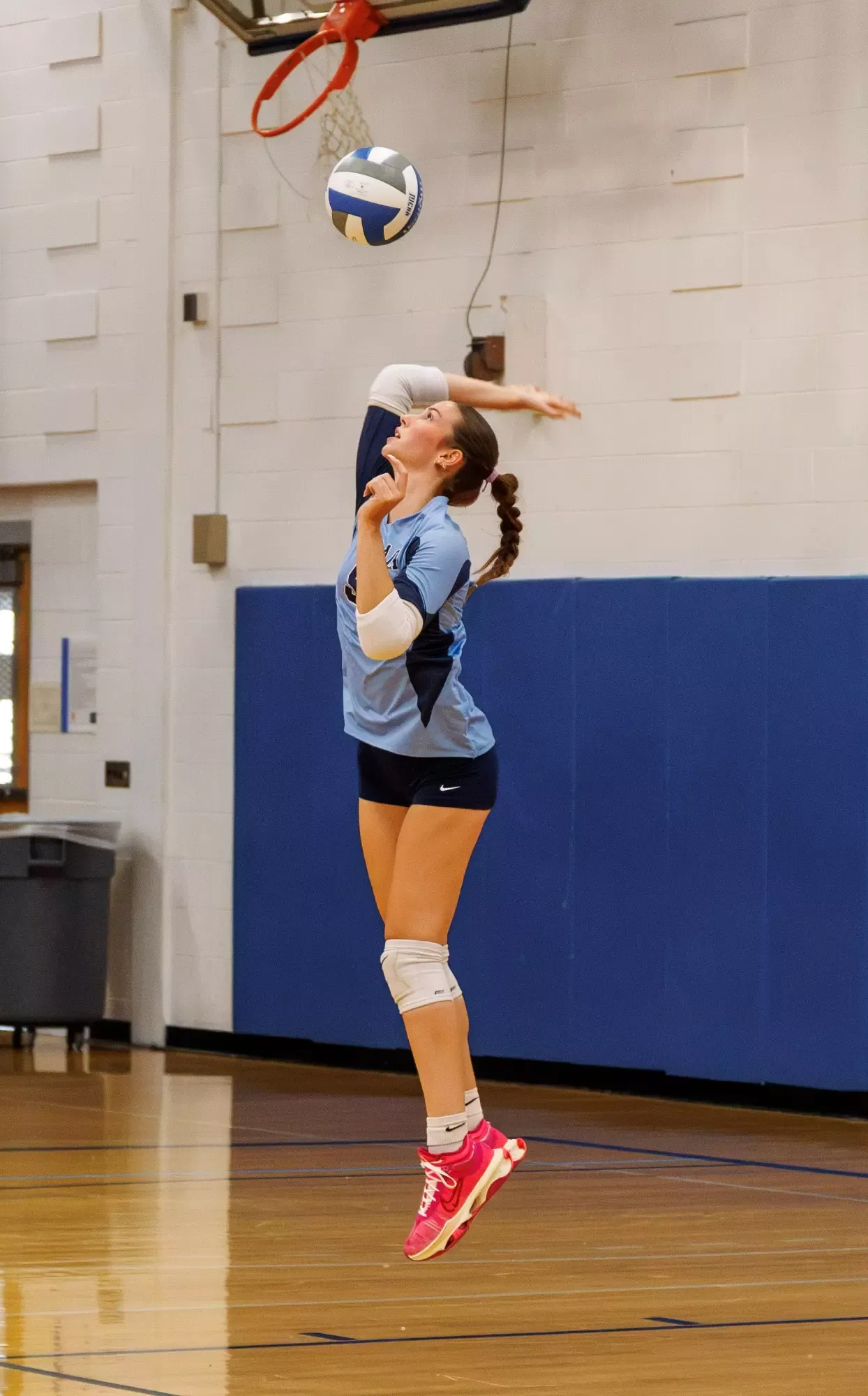 A female volleyball player is captured midair in a gymnasium, appearing to spike or serve a volleyball. She wears a light-blue jersey, black shorts, white knee pads, and bright pink athletic shoes. A volleyball net and hoop are visible above her, with blue wall padding and a polished wooden court in the background.