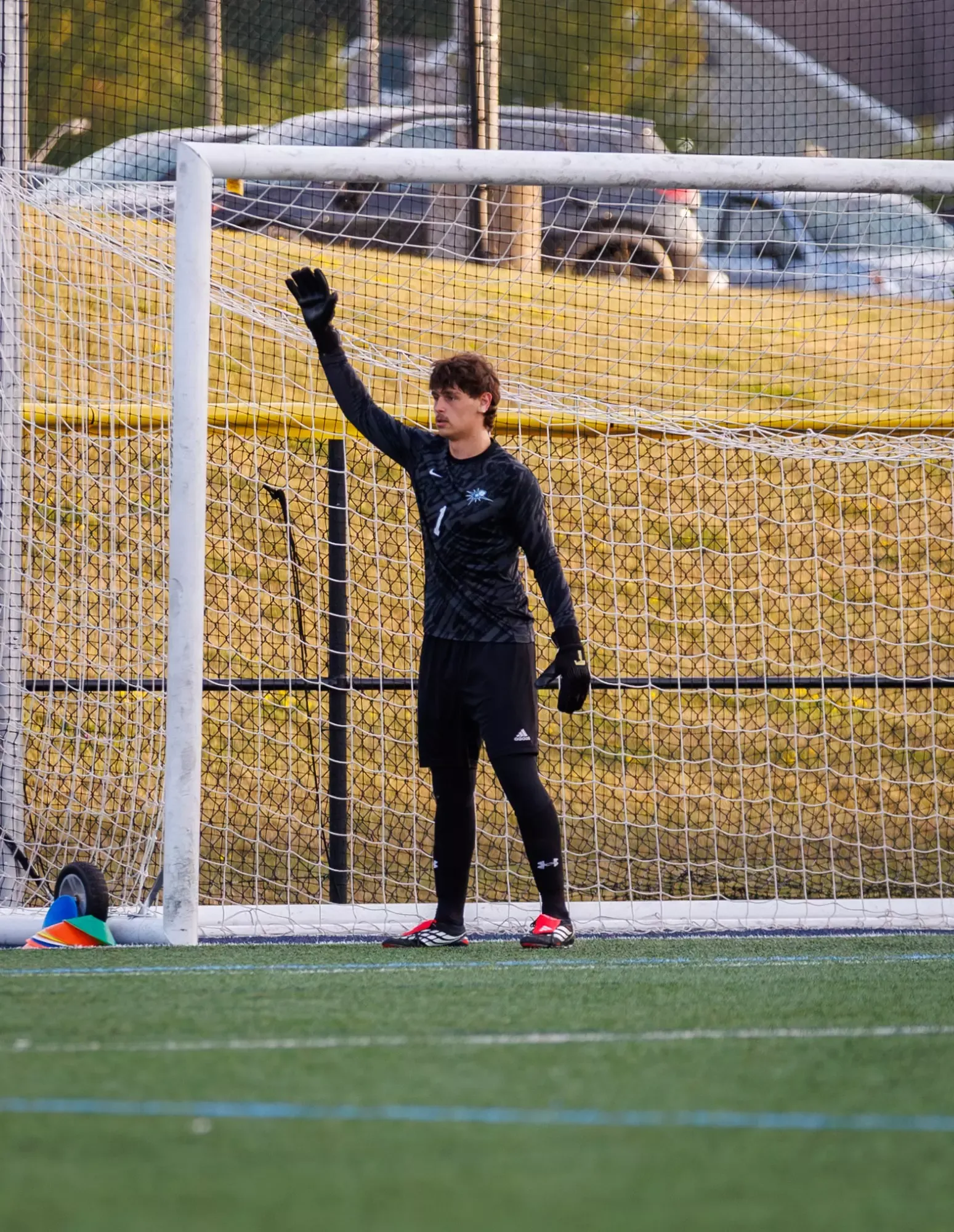 A young soccer goalkeeper in black training gear and gloves stands in front of a goal, raising one hand as he looks toward the field during practice.