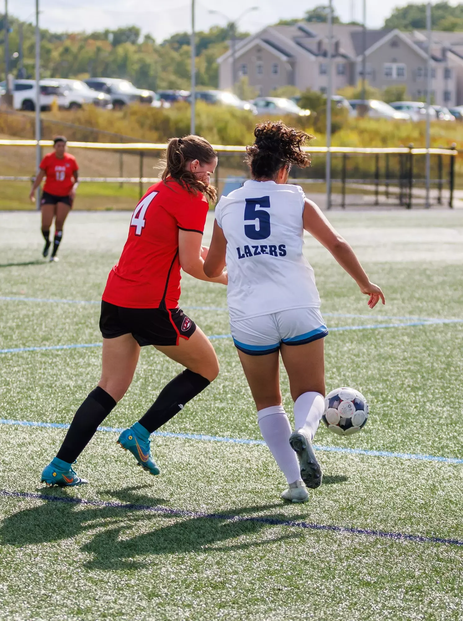 Two soccer players compete for the ball on an outdoor artificial-turf field. One player wears a red jersey with number 4 and black shorts, while the other wears a white jersey with number 5 and “LAZERS” on the back, along with white shorts. 