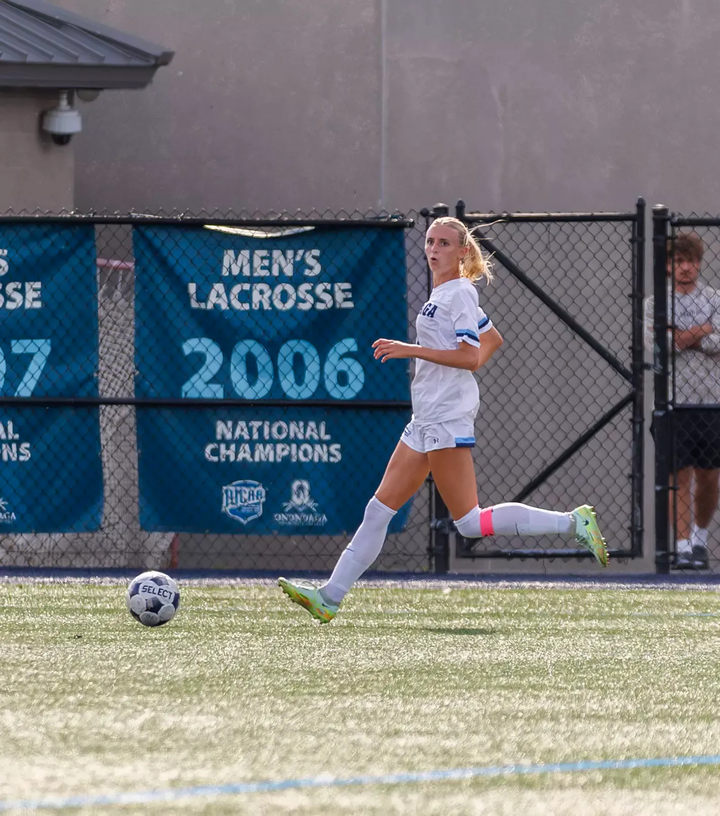 A female soccer player in a white uniform is running across a soccer field, with a soccer ball in the foreground. A chain-link fence and a large blue “MEN’S LACROSSE 2006 NATIONAL CHAMPIONS” banner are visible in the background.