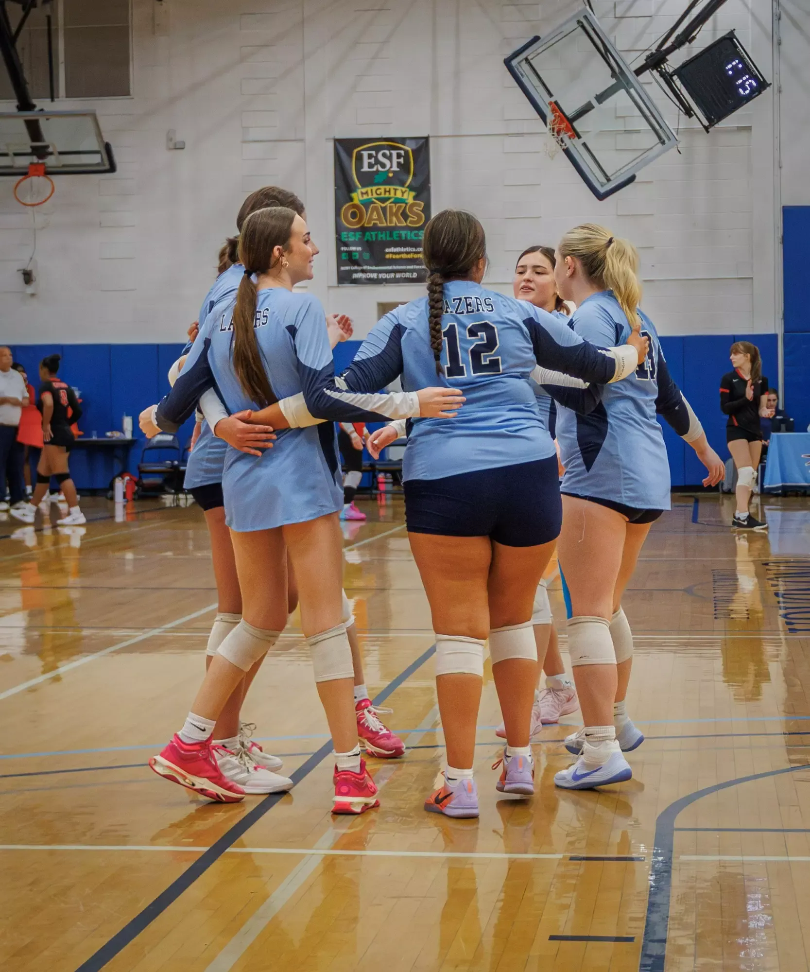 A girls’ volleyball team huddles together on an indoor basketball court during a game, with players wearing light-blue uniforms and knee pads. A scoreboard and “ESF Mighty Oaks” banner are visible in the background.